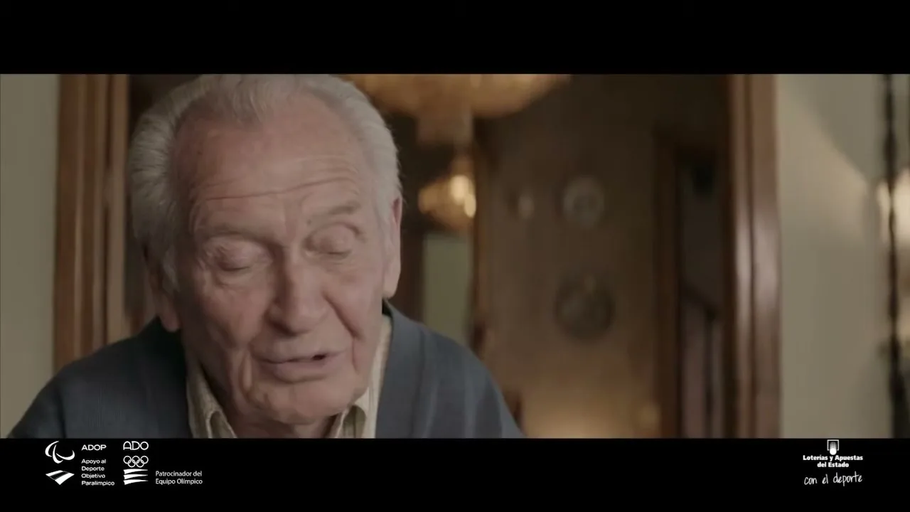An elderly man with white hair and a blue cardigan speaks directly to the camera in a softly lit interior setting.