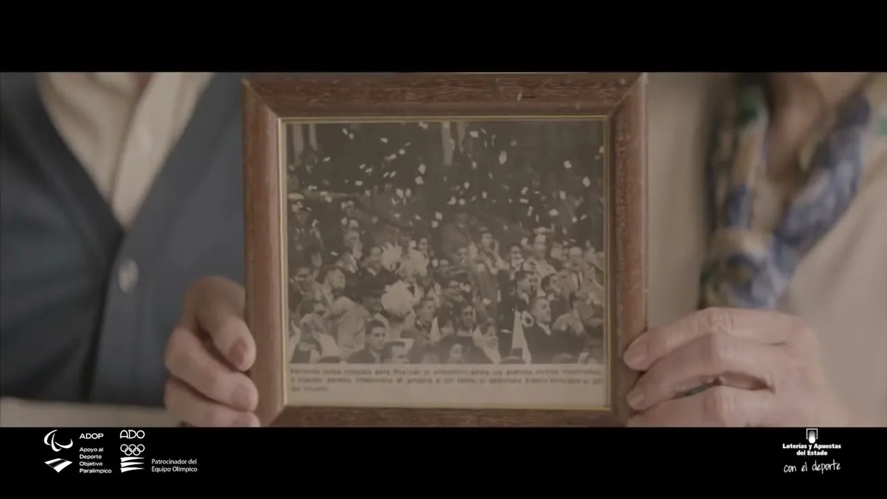 Two older people hold up a framed black and white photo of a crowd celebrating with confetti falling, in a soft focus interior setting.