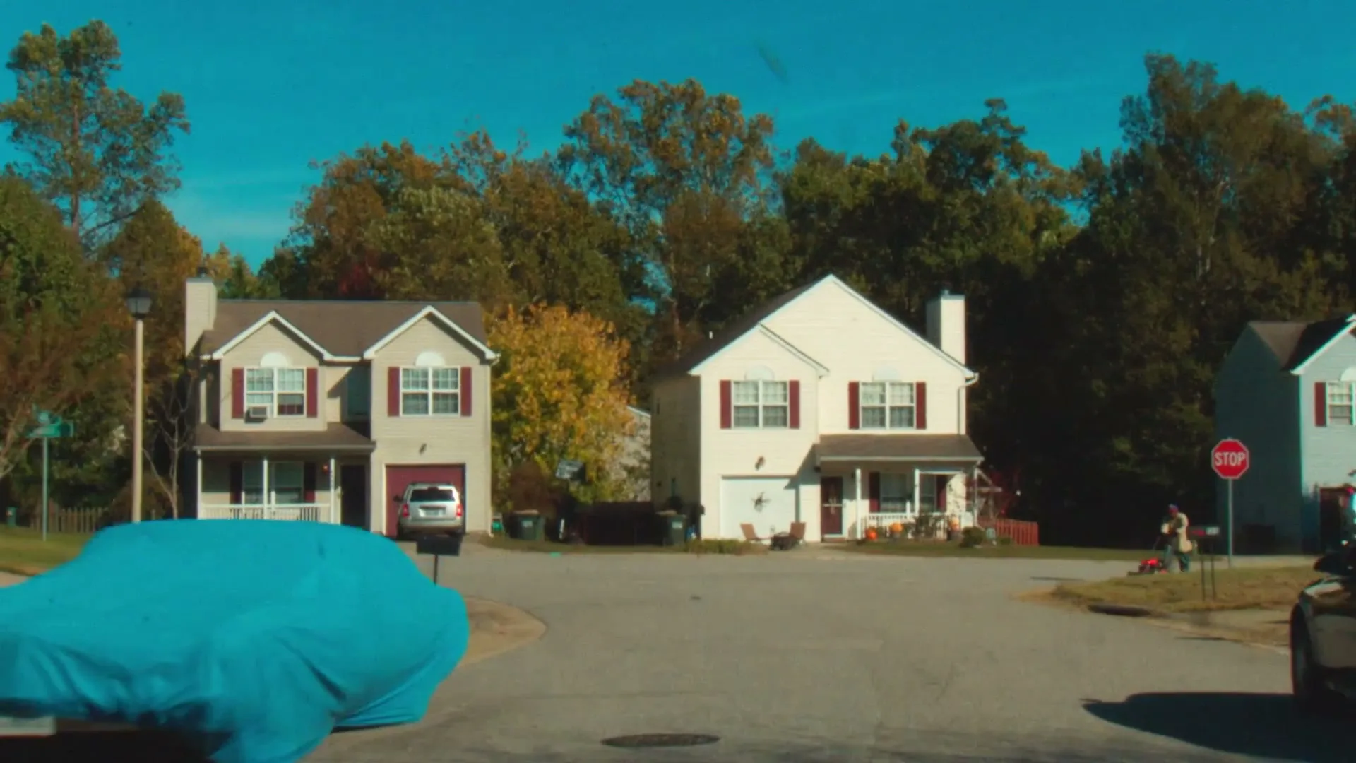 A wide shot of a suburban street shows a blue tarp covering a car, with a man mowing a lawn in the background.