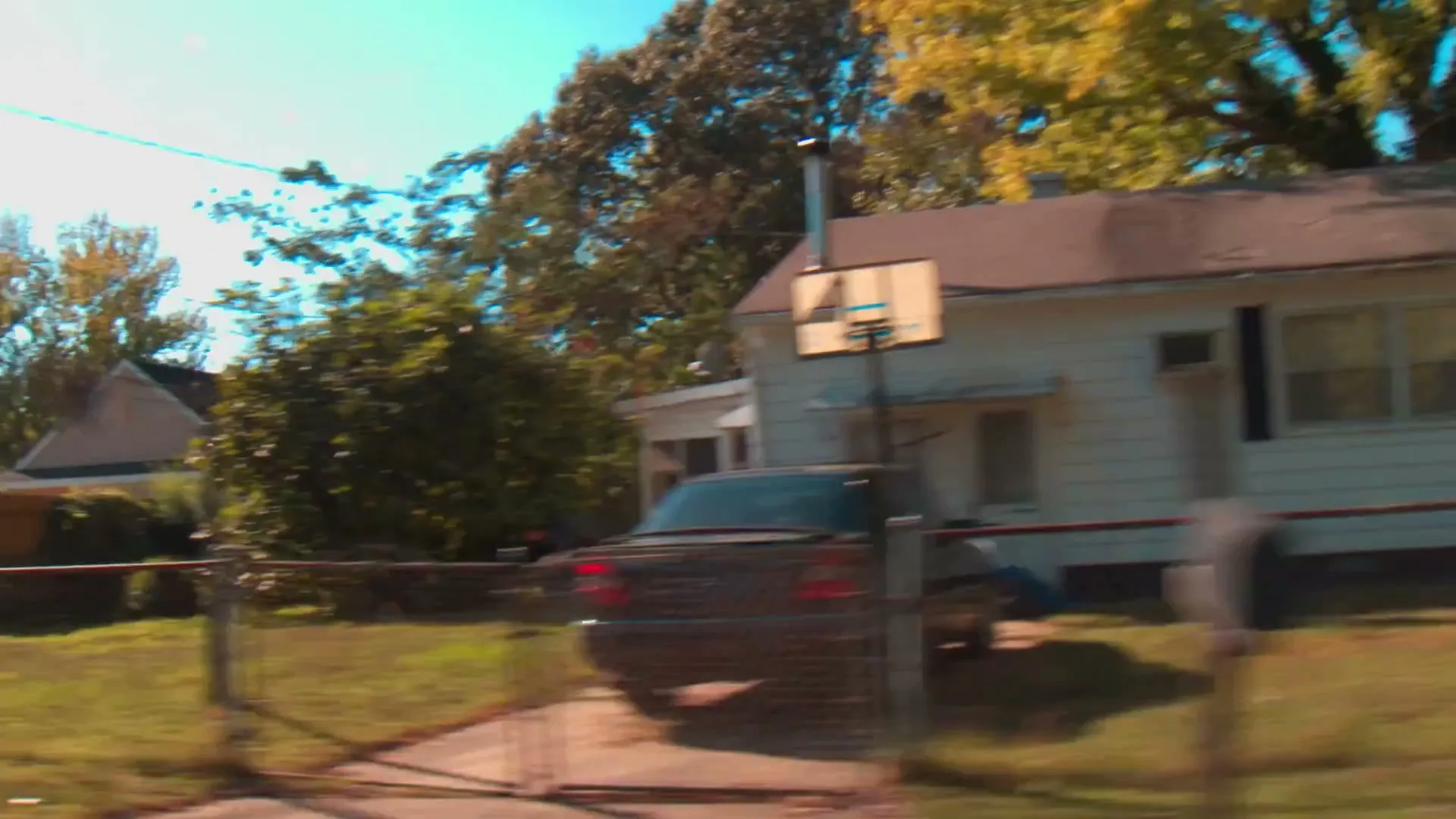A car drives past a suburban house with a basketball hoop in the driveway on a sunny day, with trees and other houses visible in the background.