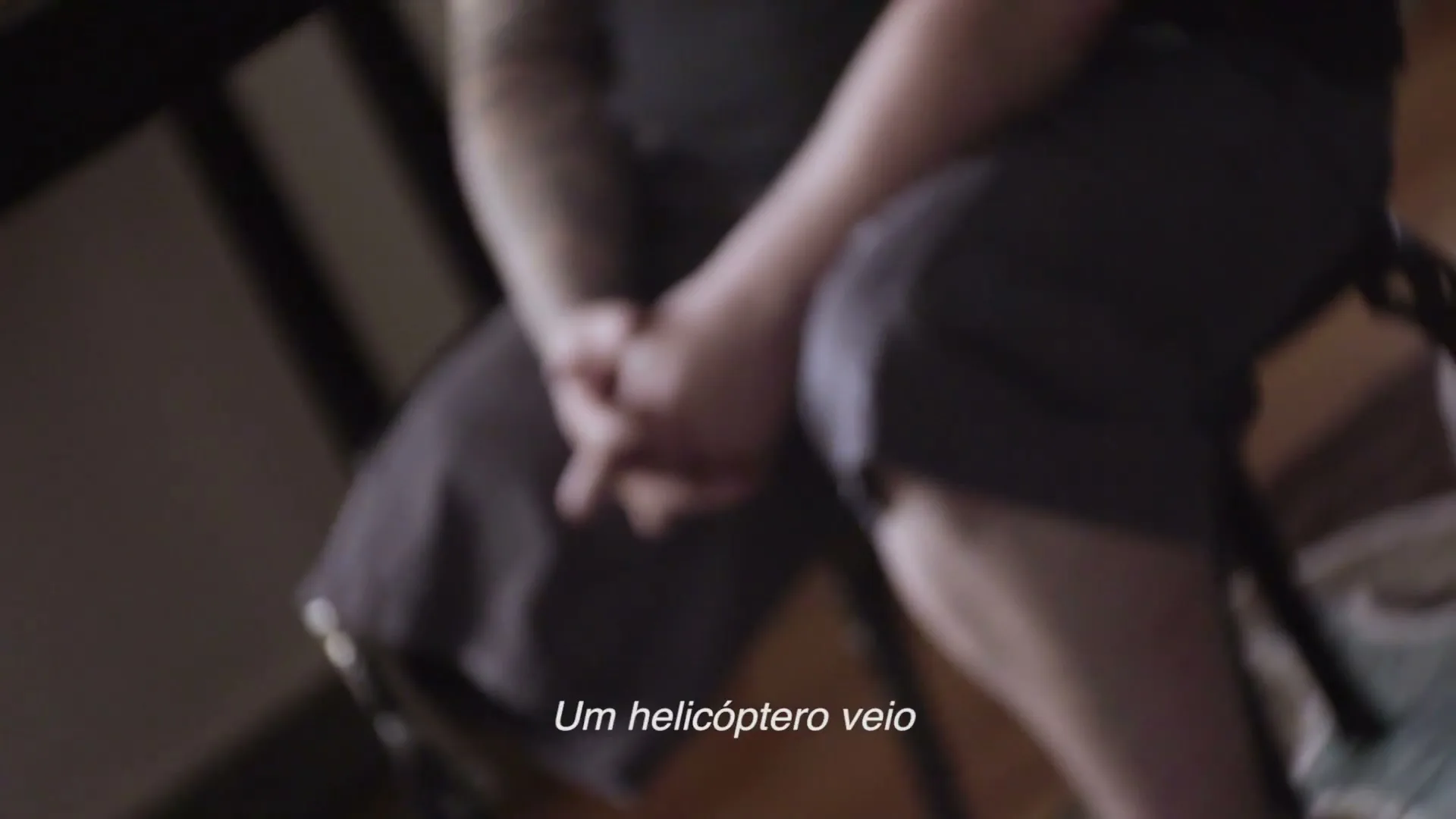 A man with tattoos on his arm sits with his hands clasped, wearing a dark t-shirt and shorts, in a dimly lit room.