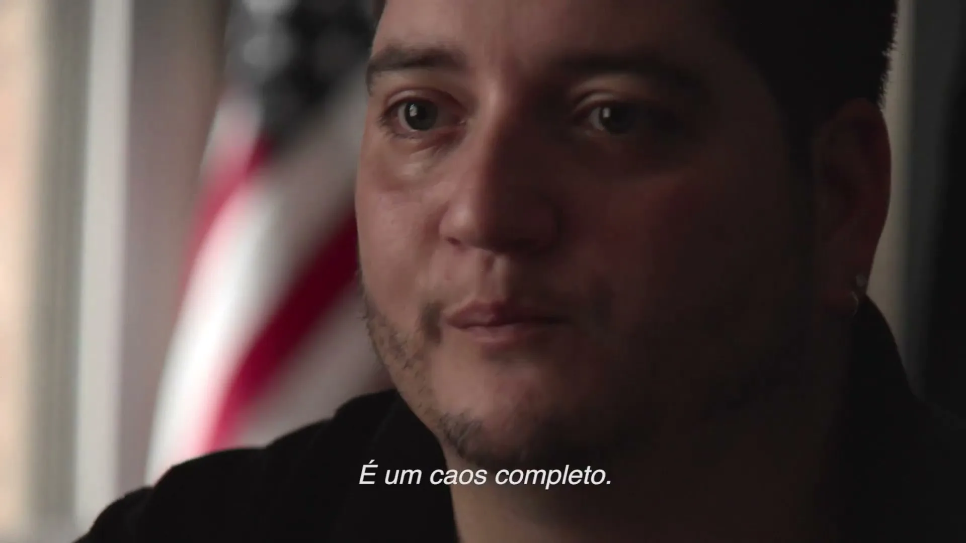 A man in his 30s with dark hair and a beard speaks to the camera, with an American flag visible in the background.