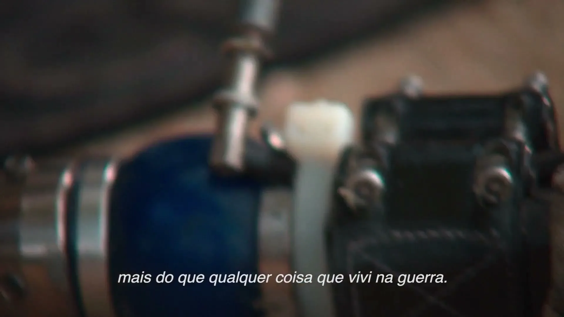 A blurry extreme closeup shot of a prosthetic leg's mechanical parts, with Portuguese subtitles at the bottom of the screen.