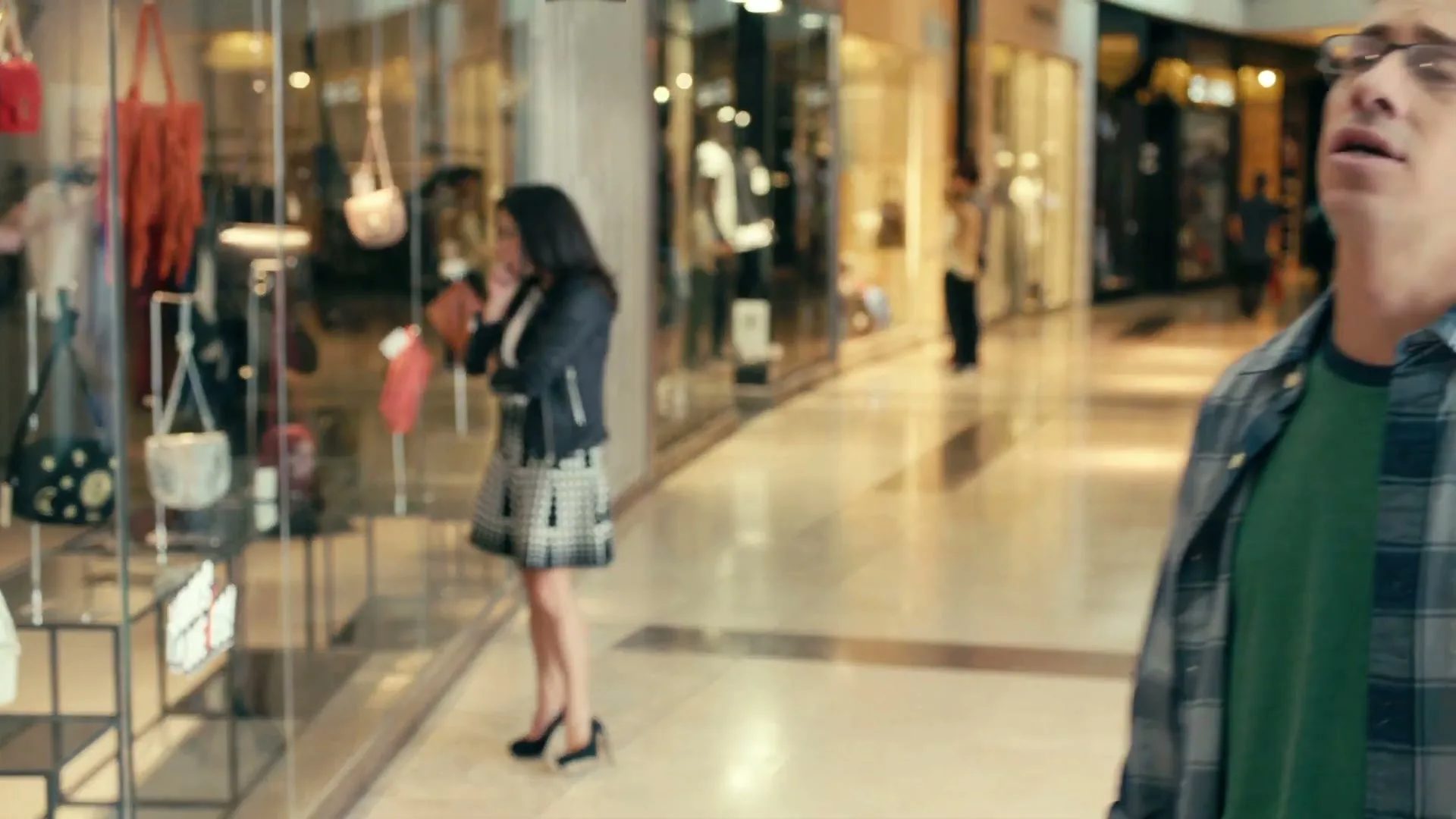 A woman in a black leather jacket and polka dot skirt looks at handbags in a store window, while a man in glasses and a plaid shirt walks past her in a shopping mall.