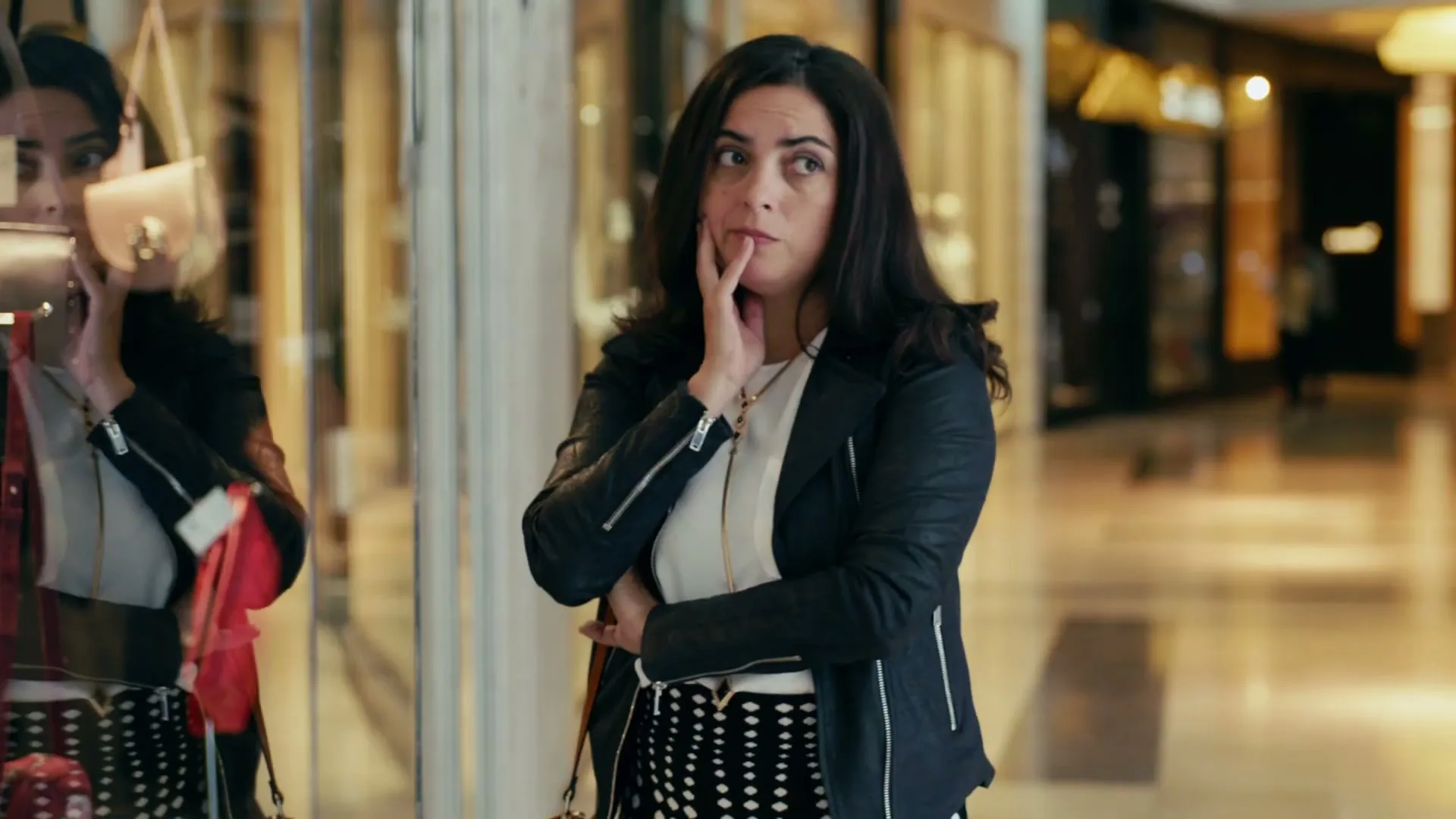 A woman with dark hair, wearing a black leather jacket and a patterned skirt, looks thoughtfully at a store window in a brightly lit shopping mall.