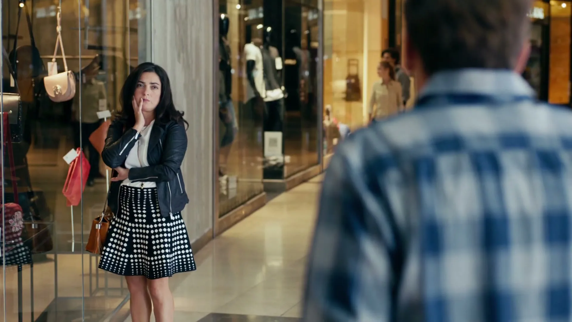 A woman in a black and white skirt looks thoughtfully at a man's back in a brightly lit shopping mall.