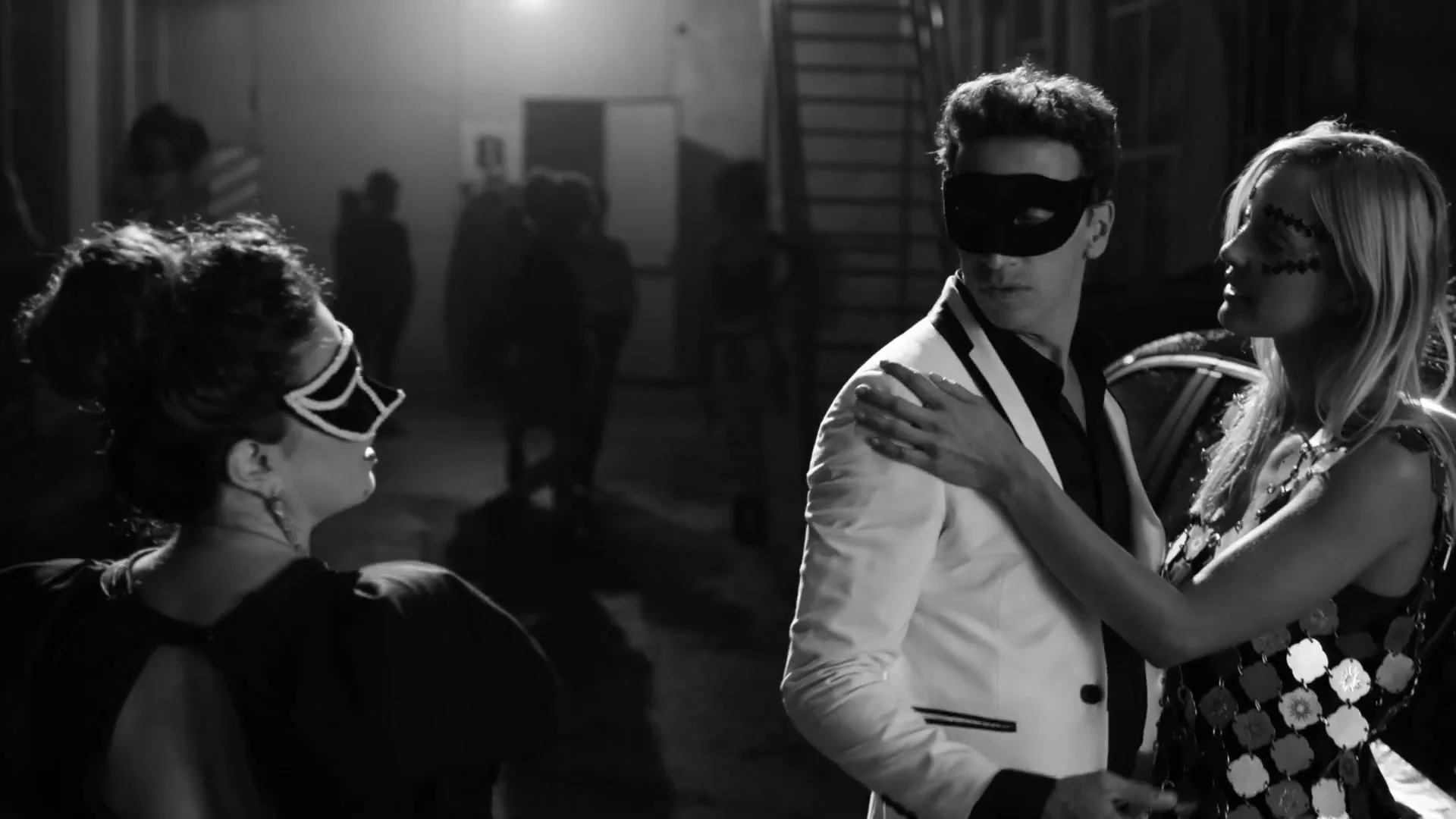 A man and woman in masquerade masks embrace at a party, while another woman in a mask looks on in a black and white shot.
