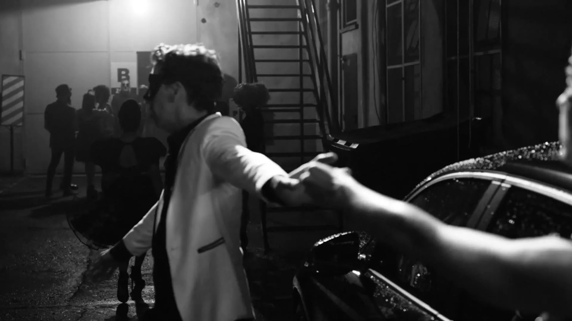 A man in a white jacket and black mask holds a woman's hand as they walk away from a party at night.