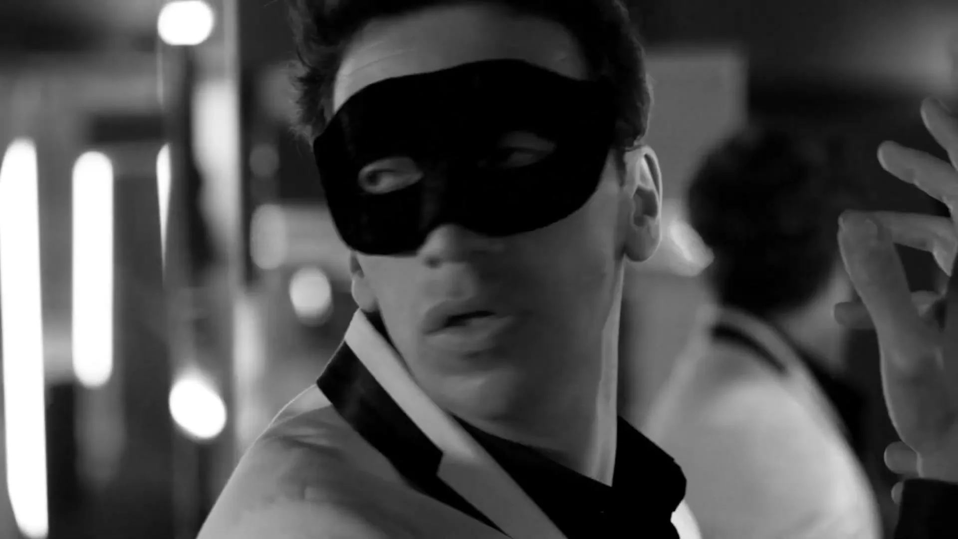 A man in a black mask and white jacket looks around with a surprised expression in a dimly lit interior.