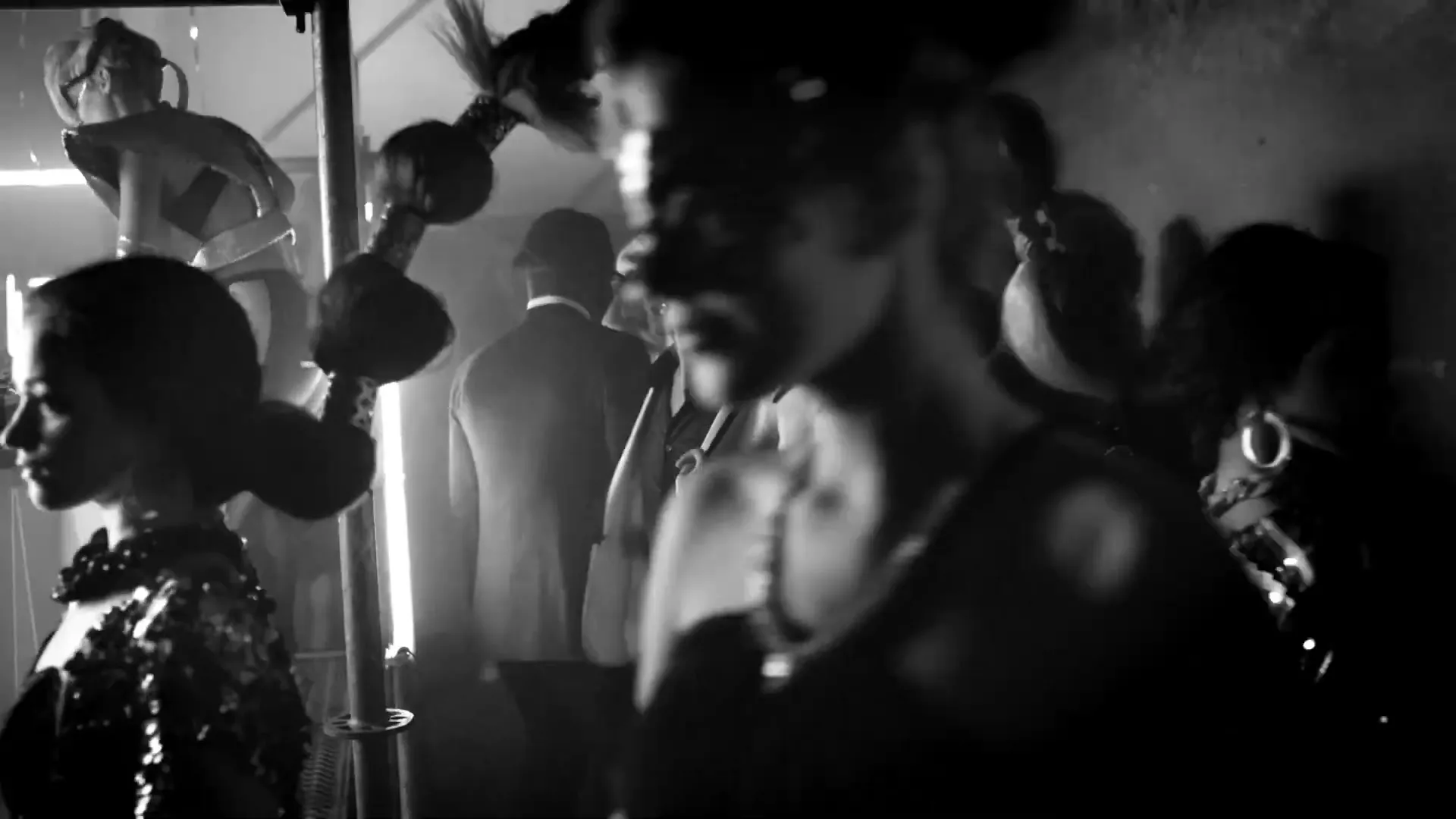 A group of people in formal wear and masks mingle at a party, with some dancing in the background, in a dimly lit interior.