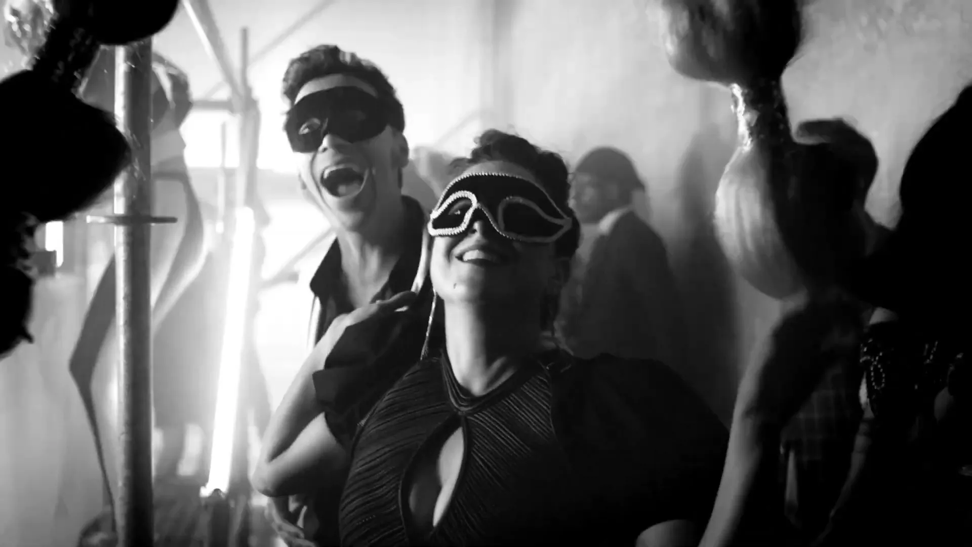 A man and a woman, both wearing masquerade masks, laugh joyfully while dancing closely together at a lively party in a dimly lit room.