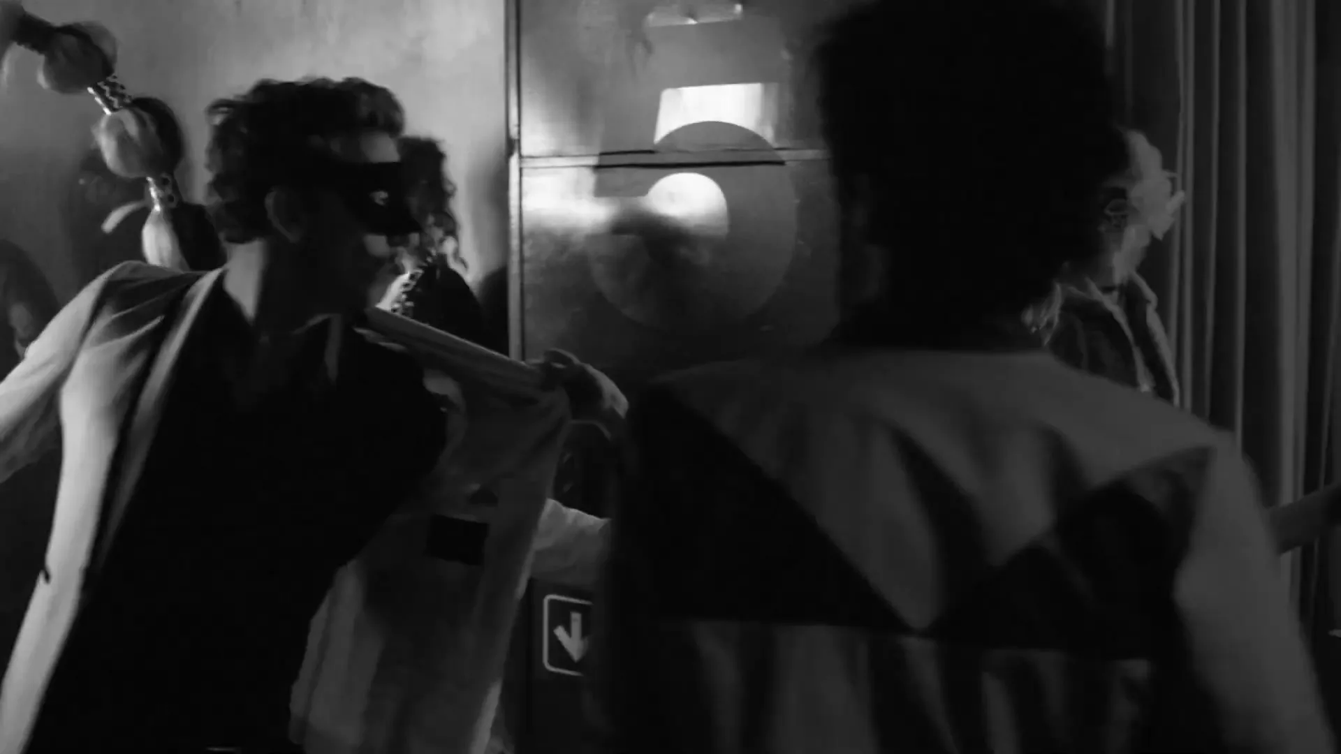 A group of people in masks and party attire are dancing and mingling in a dimly lit, crowded club, captured in black and white.