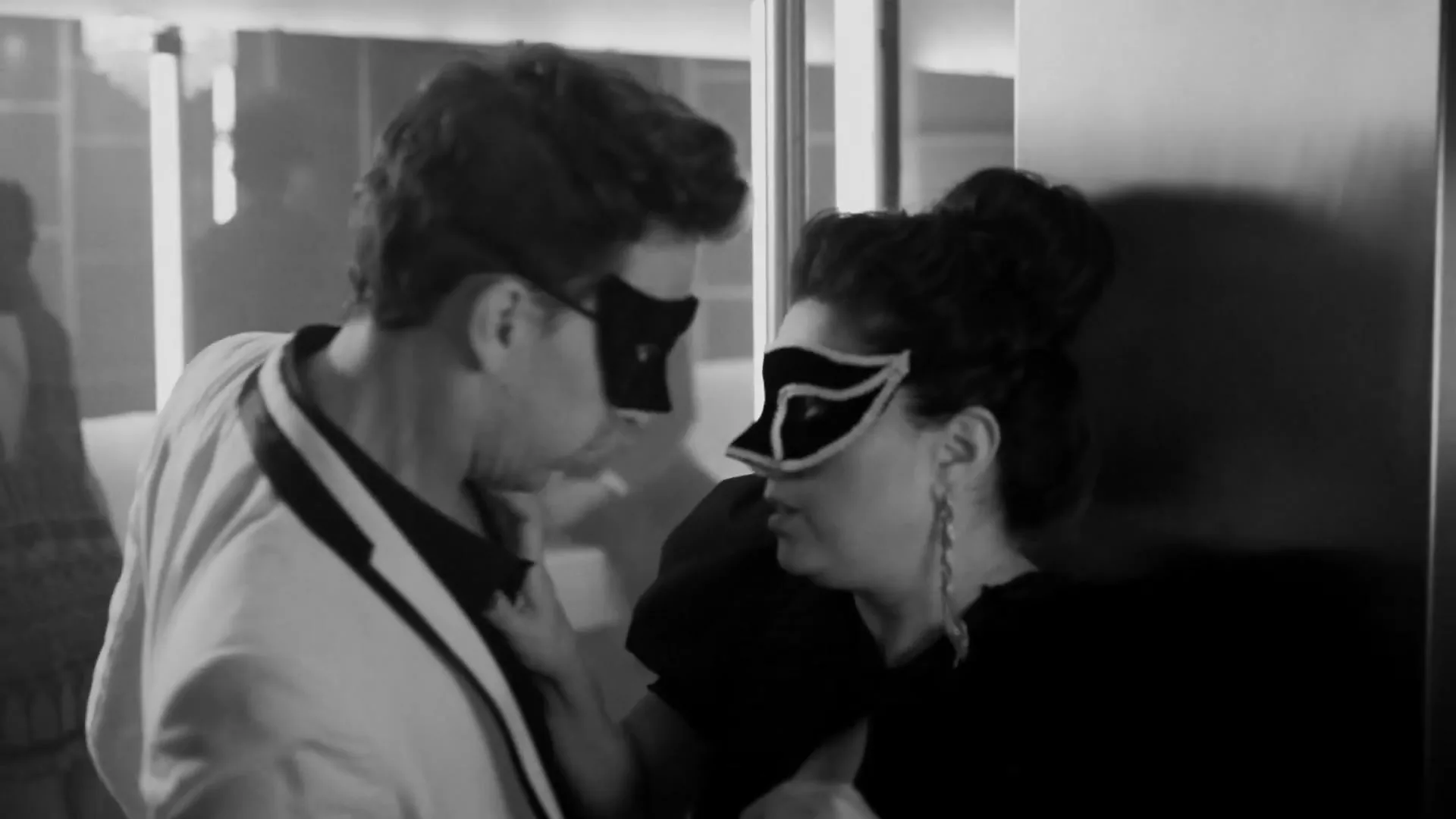 A man and a woman, both wearing masquerade masks, lean in close to each other in a dimly lit interior setting, possibly a hallway or a corner.