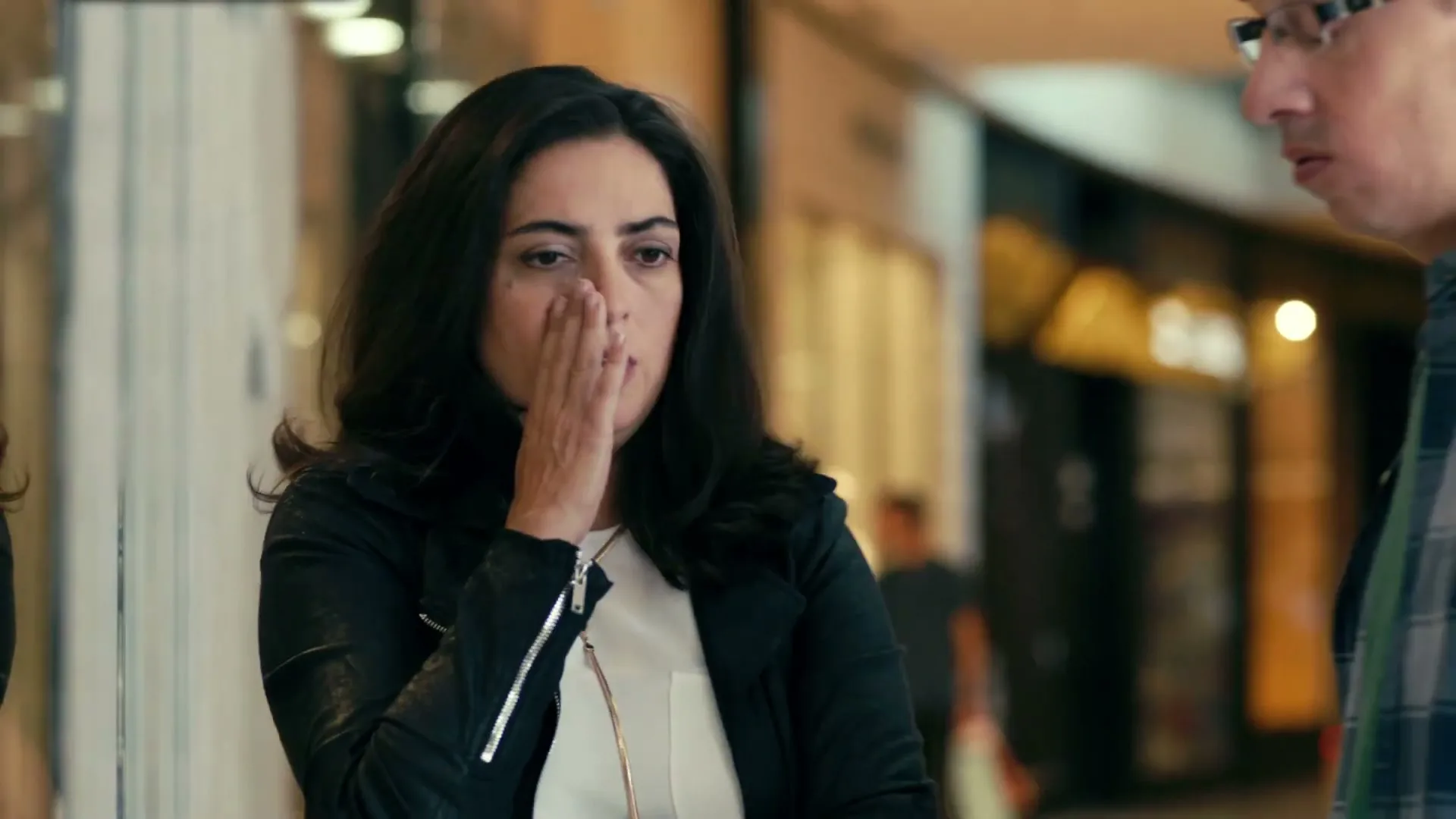A woman with dark hair and a black leather jacket looks worried and bites her nails, then covers her mouth, as a man in glasses and a plaid shirt approaches her in a mall.