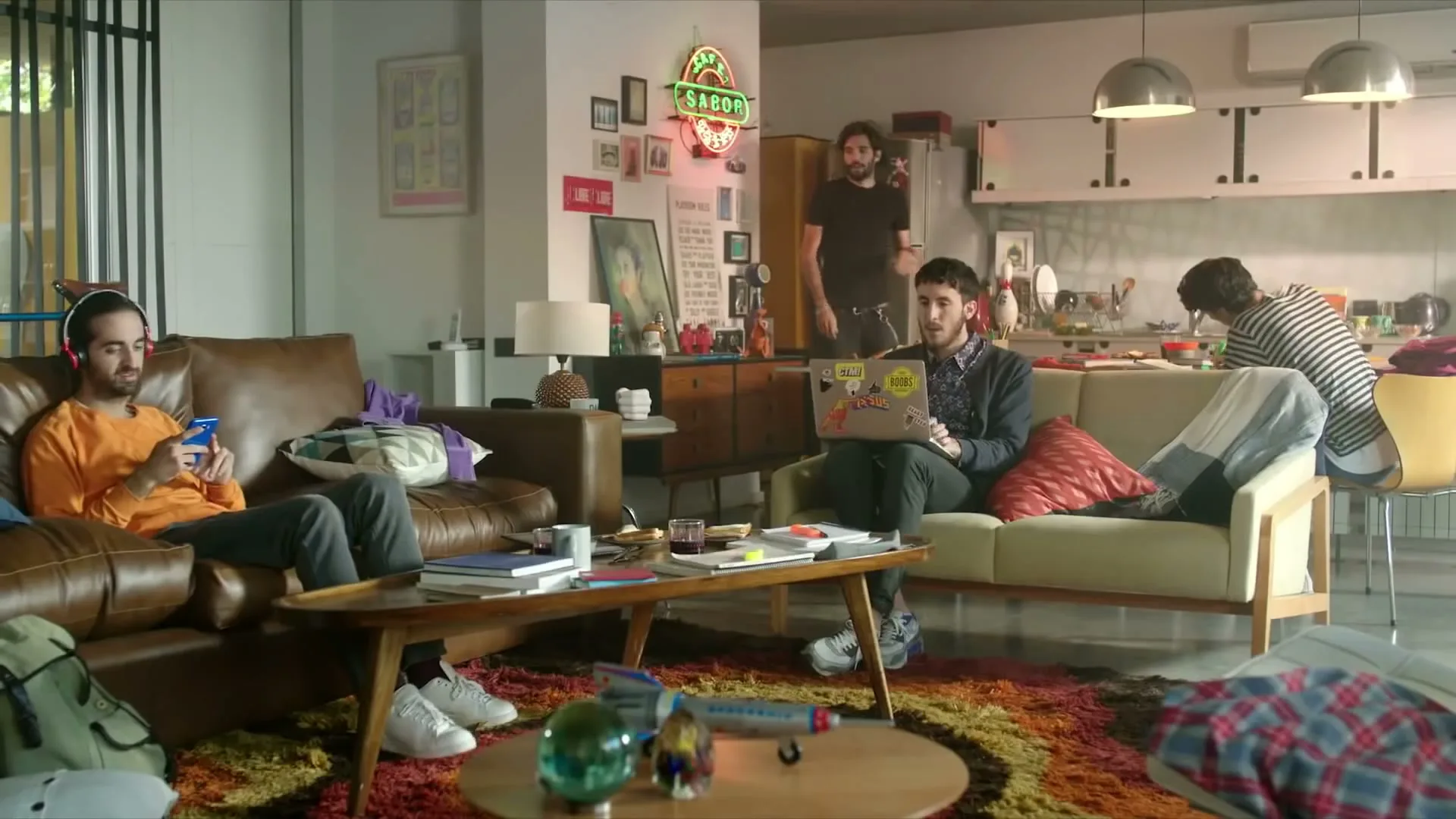 Three young men are in a messy apartment, one on a couch with headphones, another on a laptop, and a third in the kitchen.