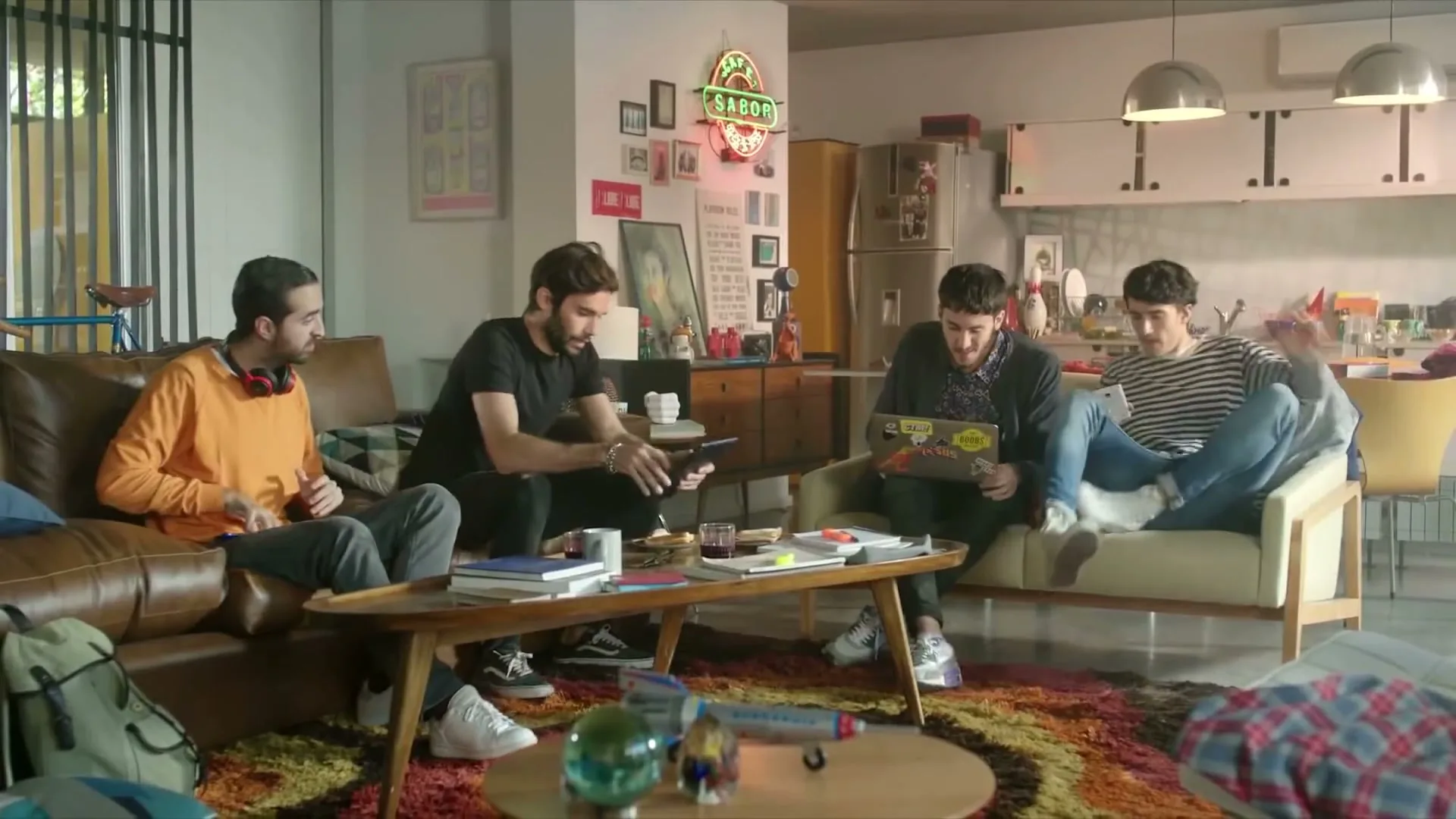 Four young men hang out in a messy living room, one playing a video game, another on a laptop, and two others chatting.