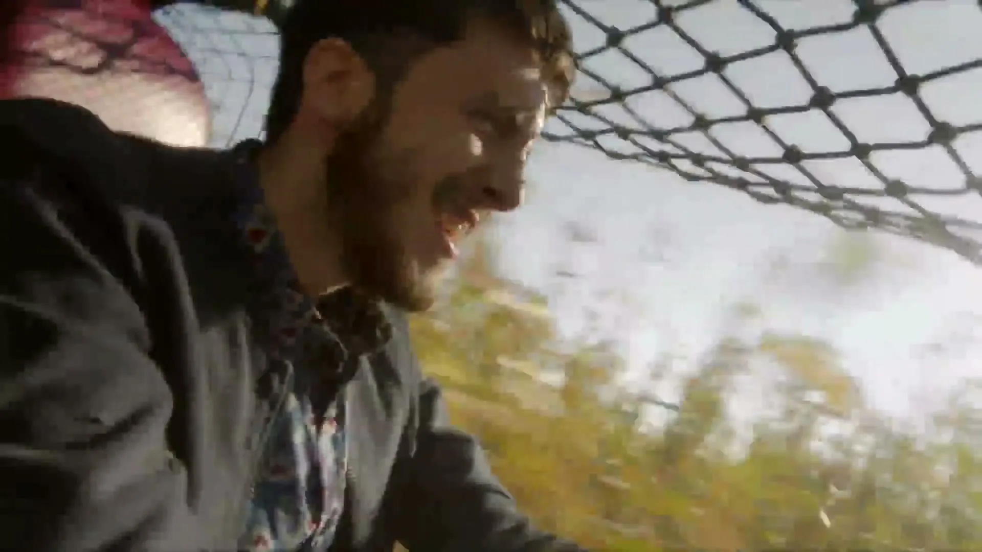 A man with a beard, wearing a dark jacket, smiles excitedly while riding in a vehicle with a cargo net overhead, speeding through a green landscape.