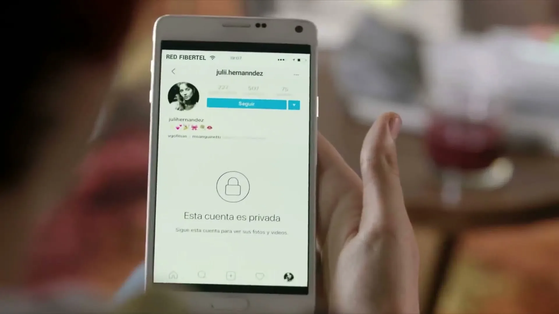 A person holds a white smartphone displaying a private Instagram profile with a lock icon and text in Spanish.