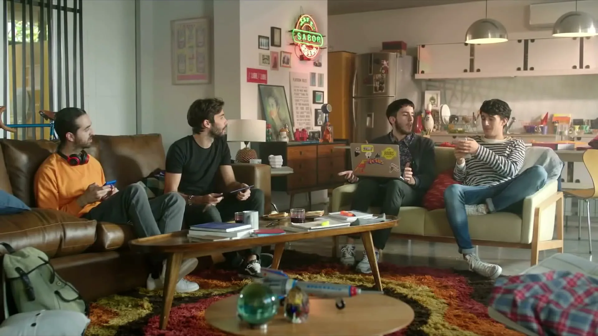 Four young men are sitting in a living room, two on a brown leather couch, one on a yellow couch with a laptop, and one on the yellow couch holding a phone.