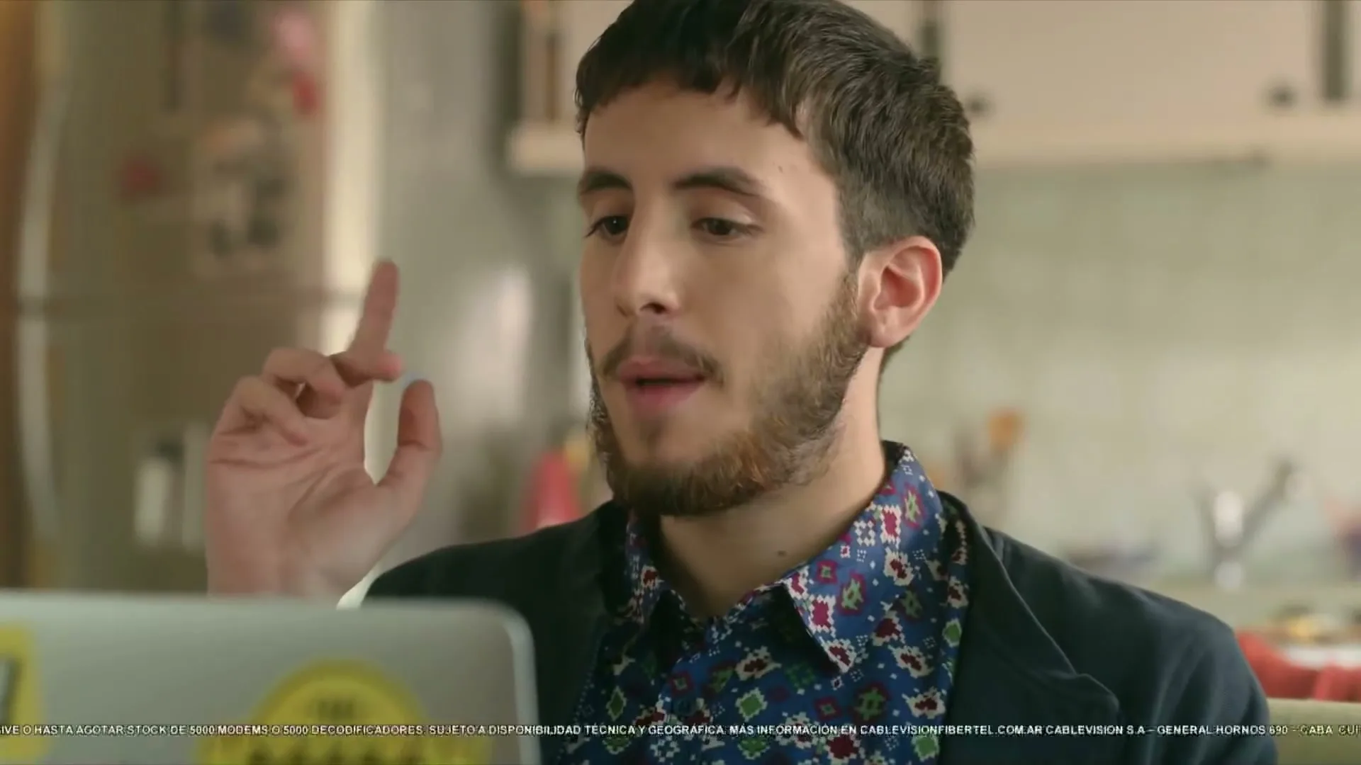 A young man with a beard and patterned shirt sits in a kitchen, looking at a laptop and gesturing with his hand.