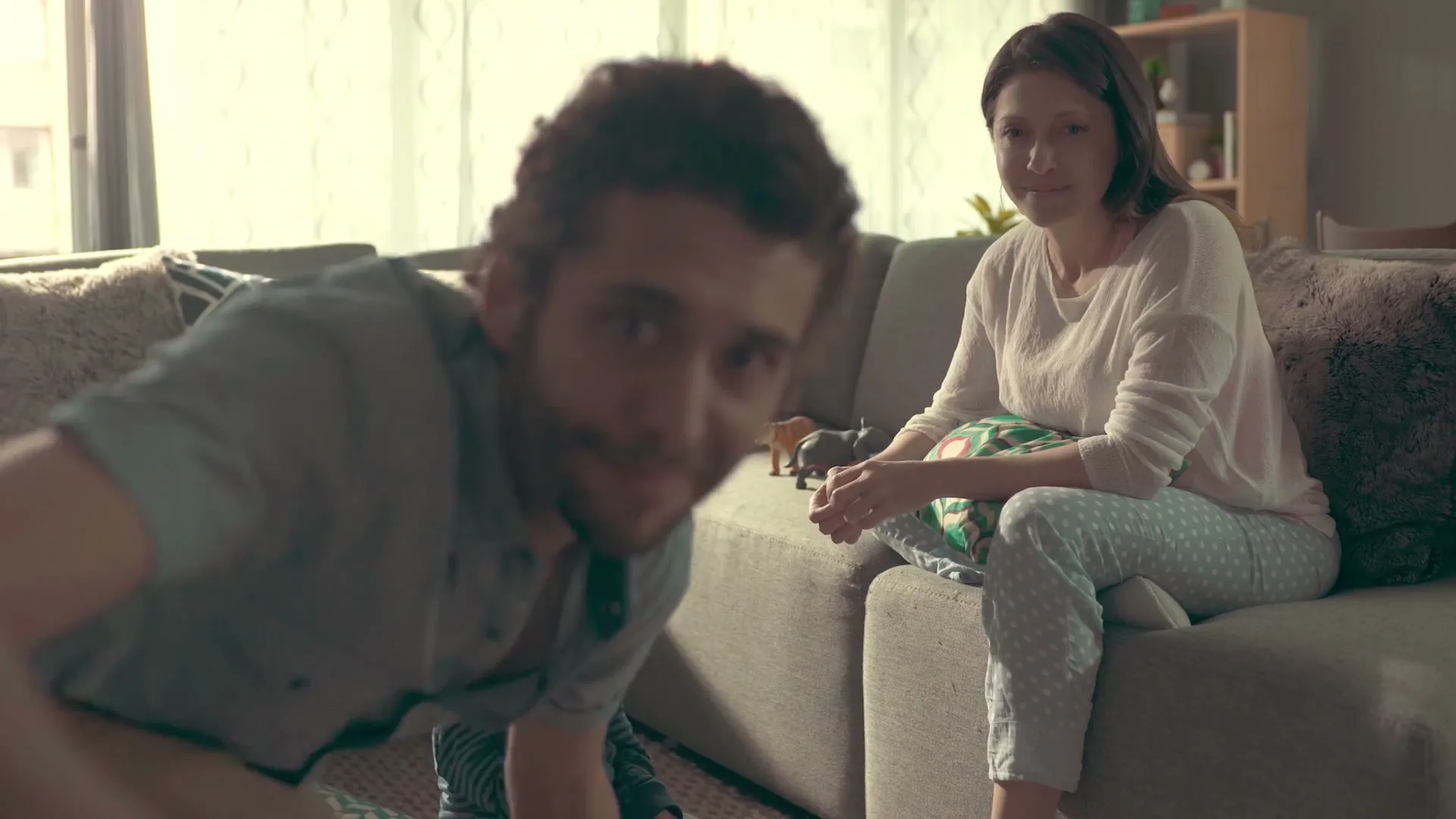 A man with a beard and a woman in pajamas are in a living room, with the man looking at the camera and the woman smiling.