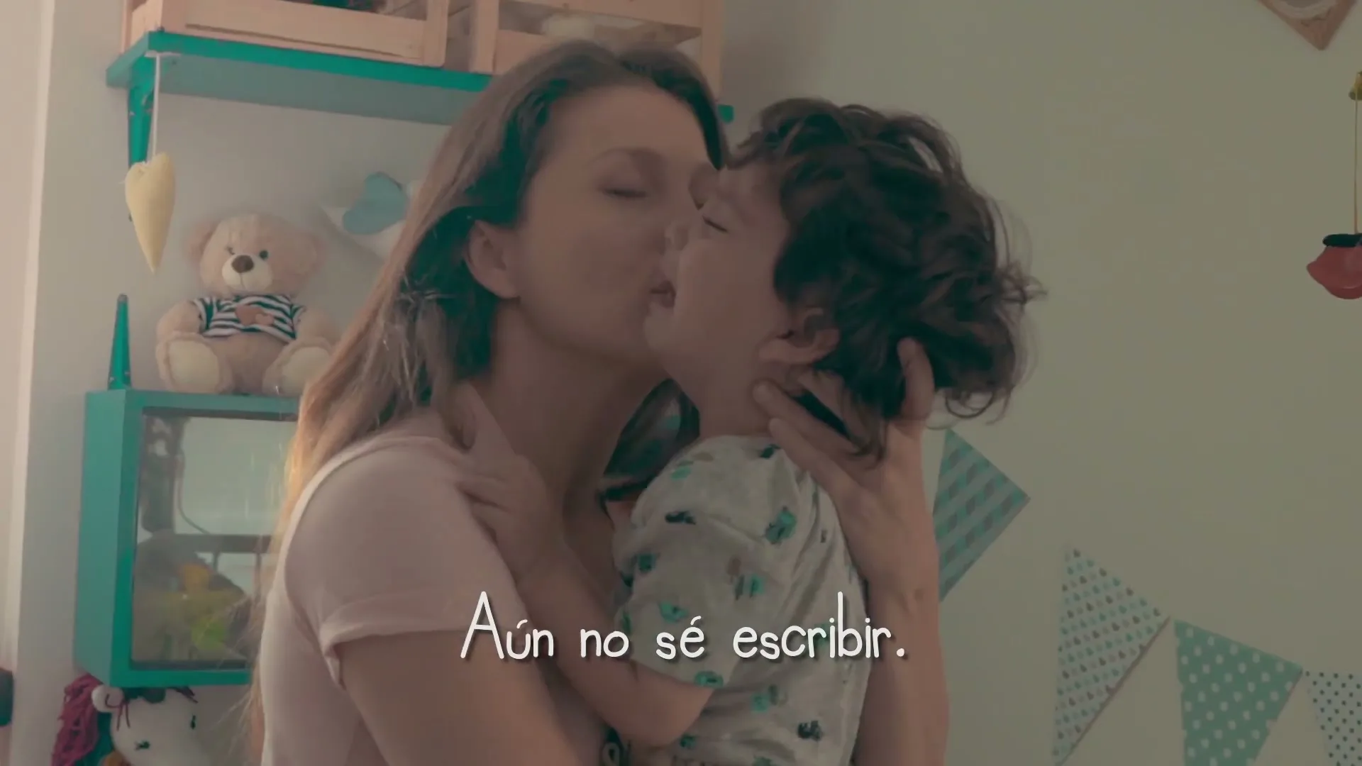 A mother with long brown hair in a pink shirt holds and kisses her young son in a brightly lit child's bedroom.