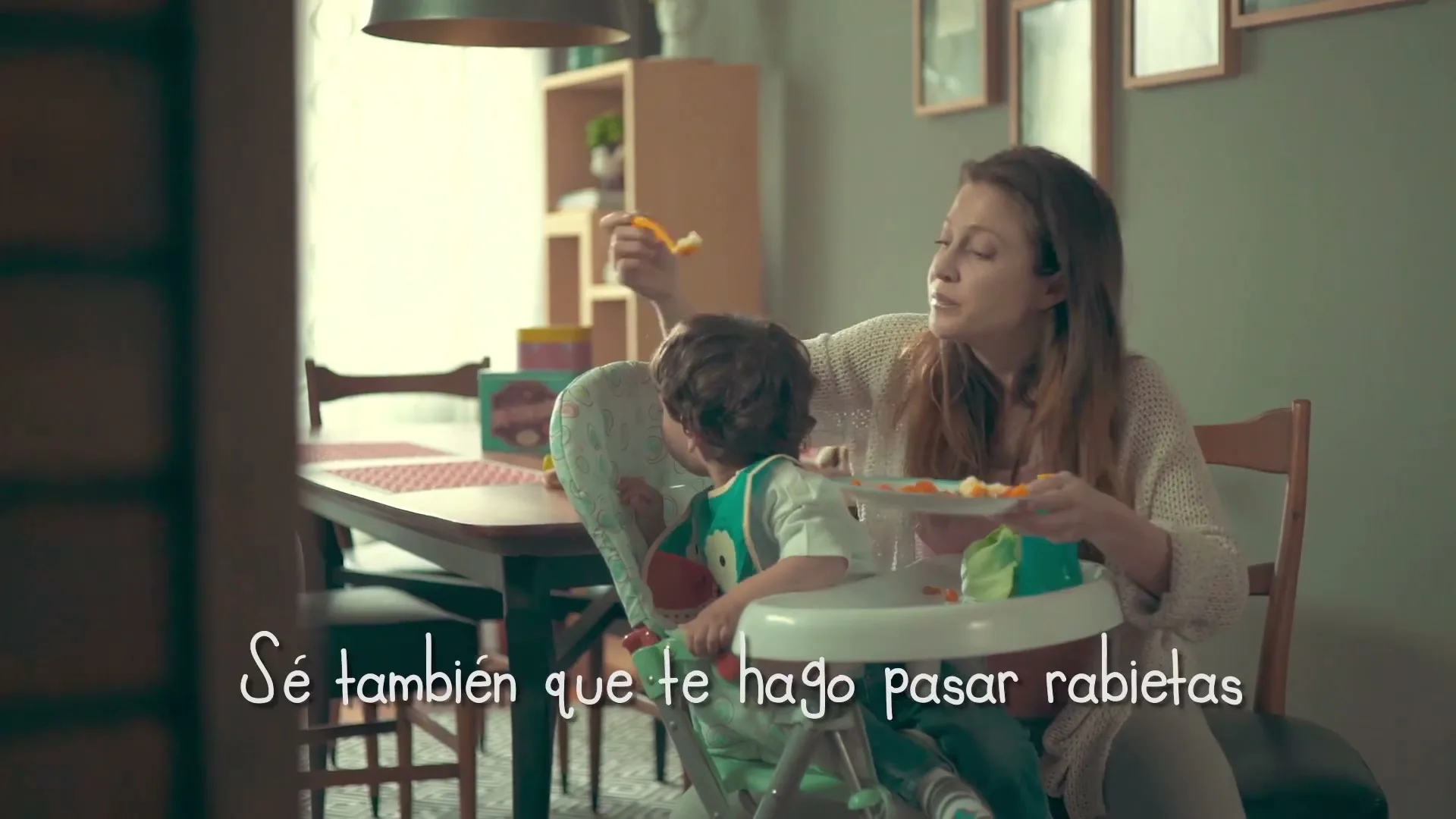 A mother with long brown hair feeds her young son in a high chair at a dining table in their home, as he looks away.