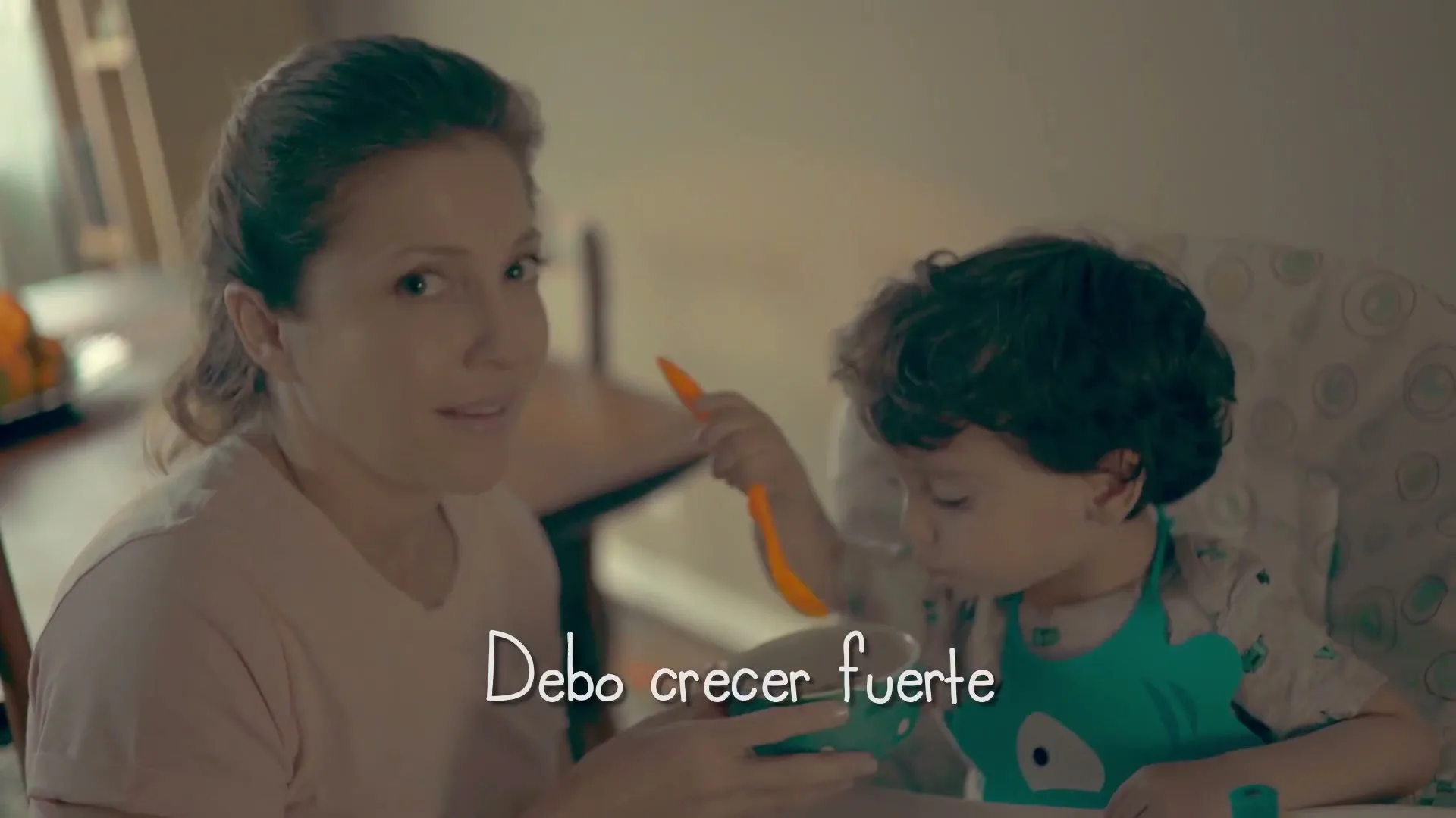 A mother with her hair tied back, wearing a light pink shirt, smiles at the camera while holding a bowl for her young son, who is eating with an orange spoon in a high chair.