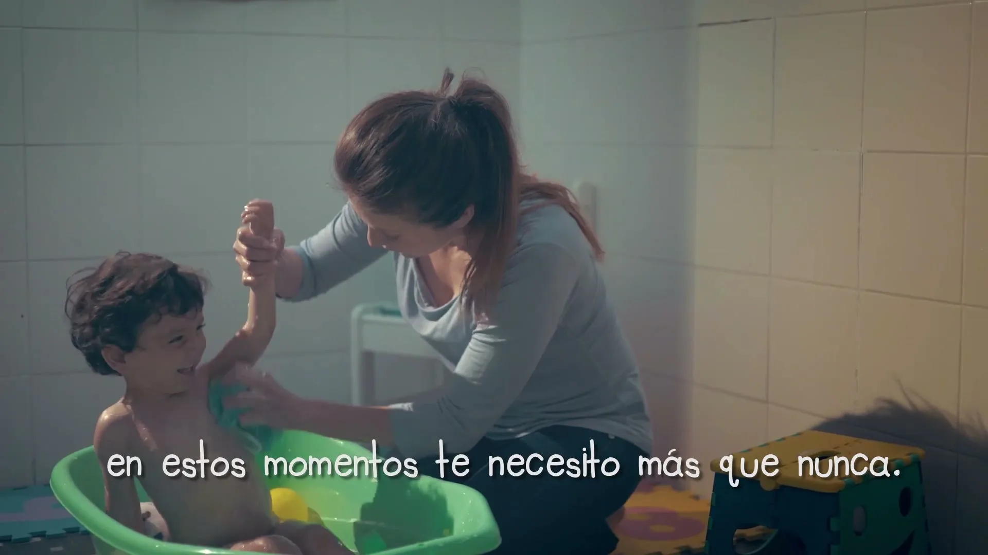 A mother with red hair in a ponytail bathes her young son in a green tub, scrubbing his arm with a sponge in a tiled bathroom.
