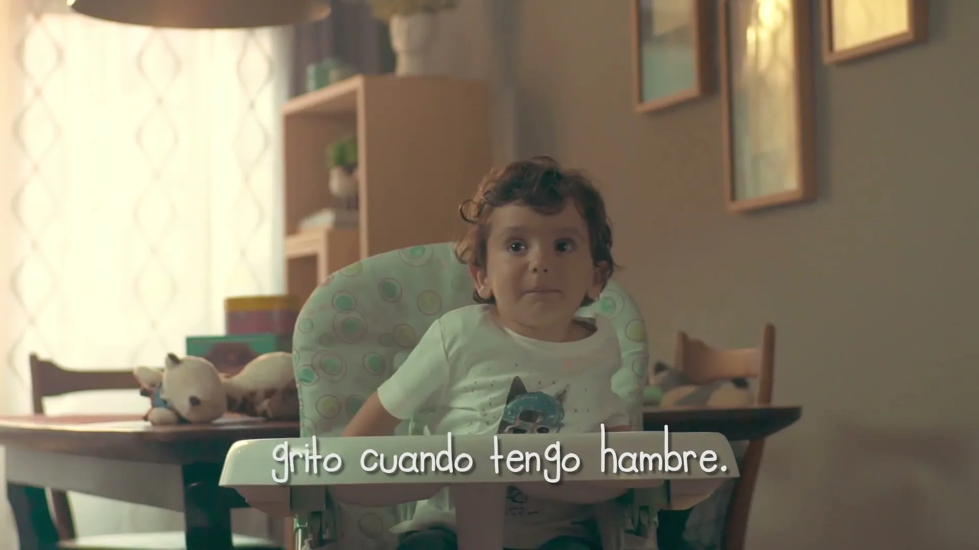 A baby with curly hair sits in a high chair, looking around a dining room with a toy giraffe on the table.