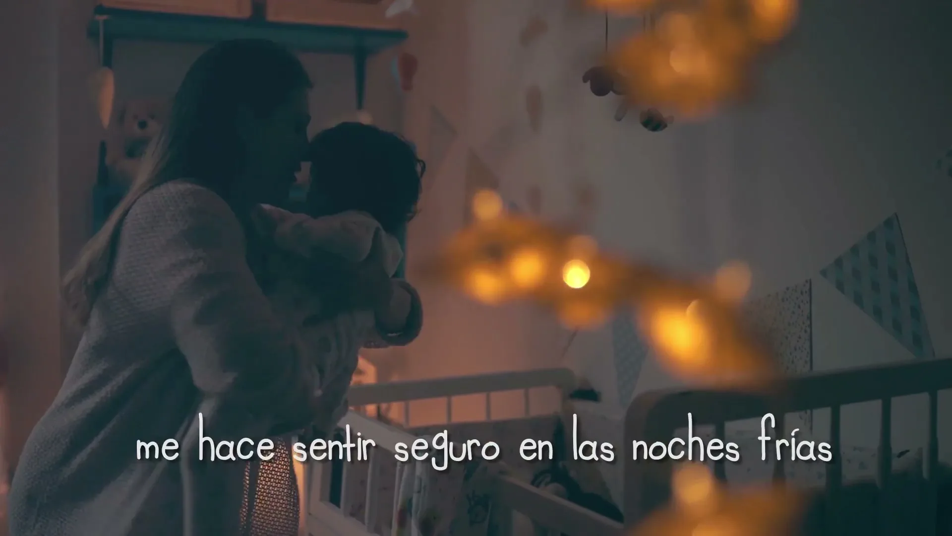 A mother in her 30s, wearing a light-colored robe, holds her baby in a dimly lit nursery, gently swaying near a crib with glowing string lights.