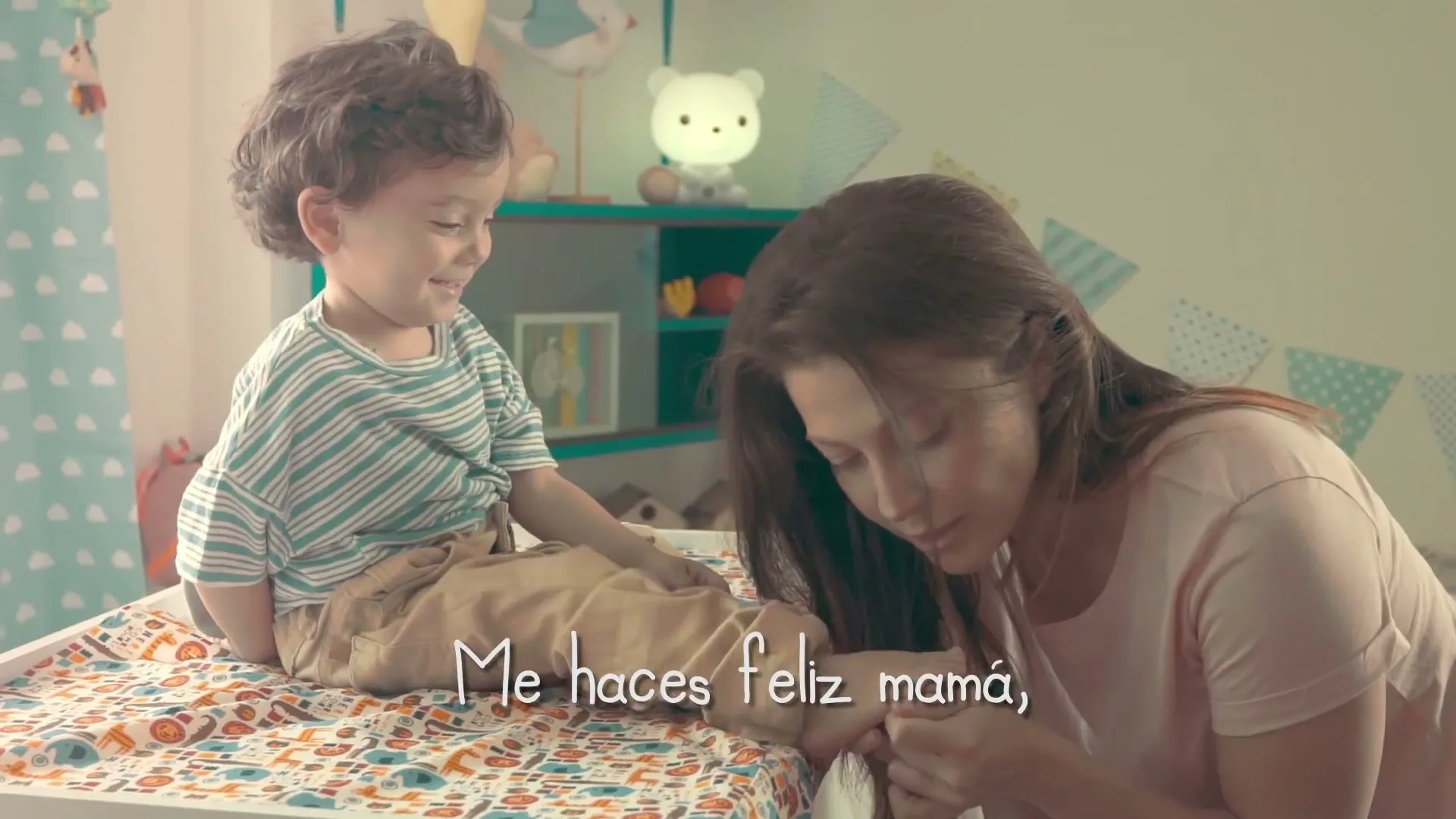 A young boy with curly hair sits on a changing table, smiling at his mother who is tickling his foot in a brightly lit nursery.