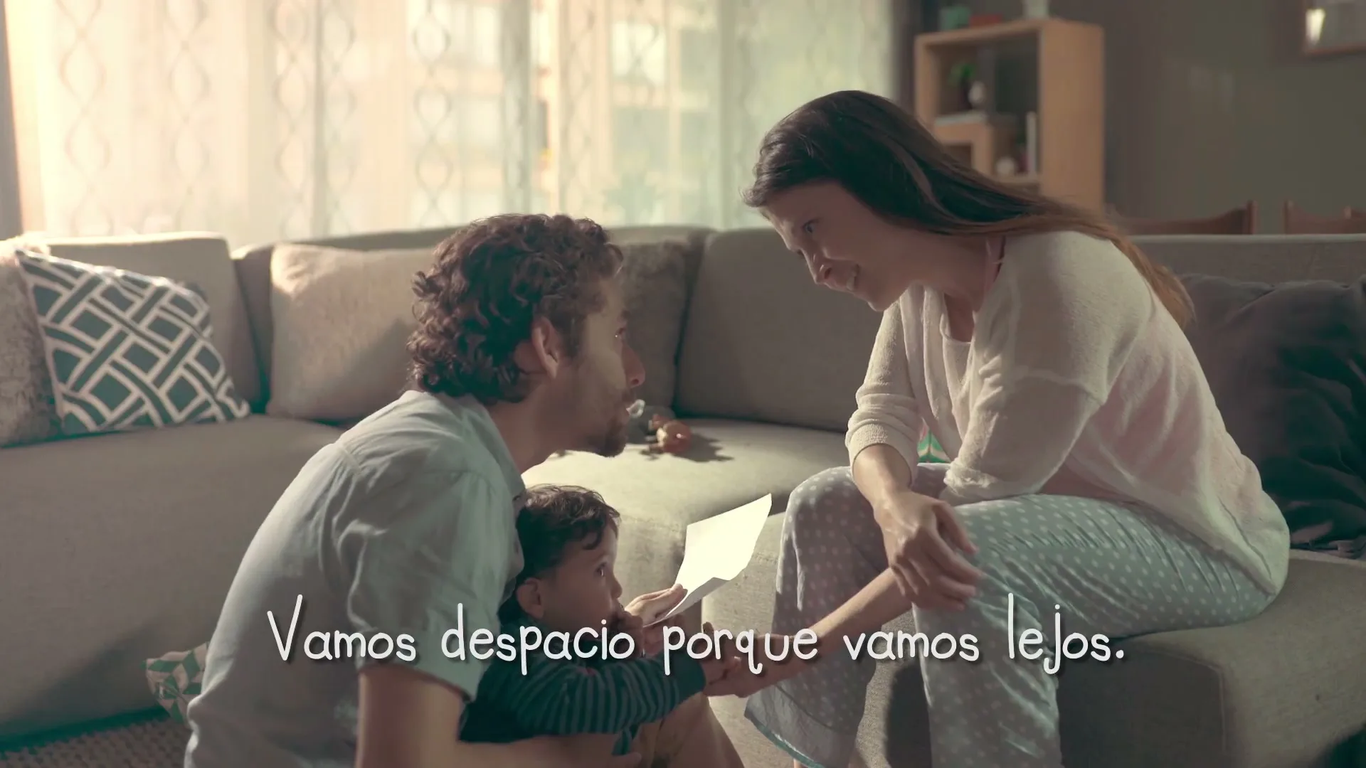 A father, mother, and baby sit on the floor and couch in a living room, looking at a letter together, with text on screen.
