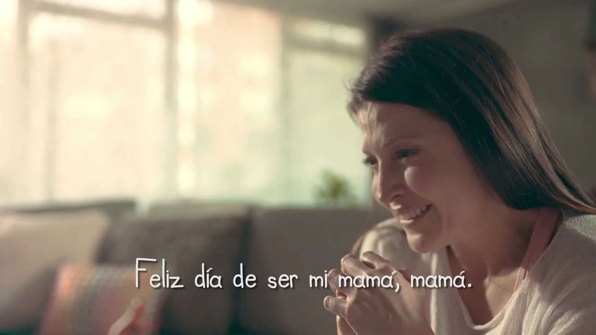 A woman with long brown hair and a pink apron smiles and looks down, with text on screen, in a living room.