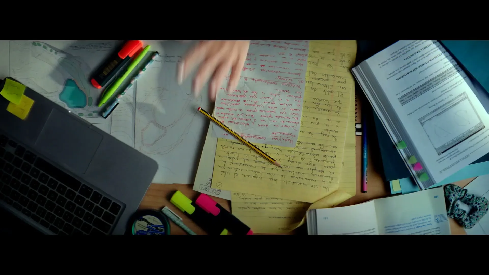 A female hand reaches for a pencil on a cluttered desk with a laptop, books, and papers, suggesting a study or work environment.