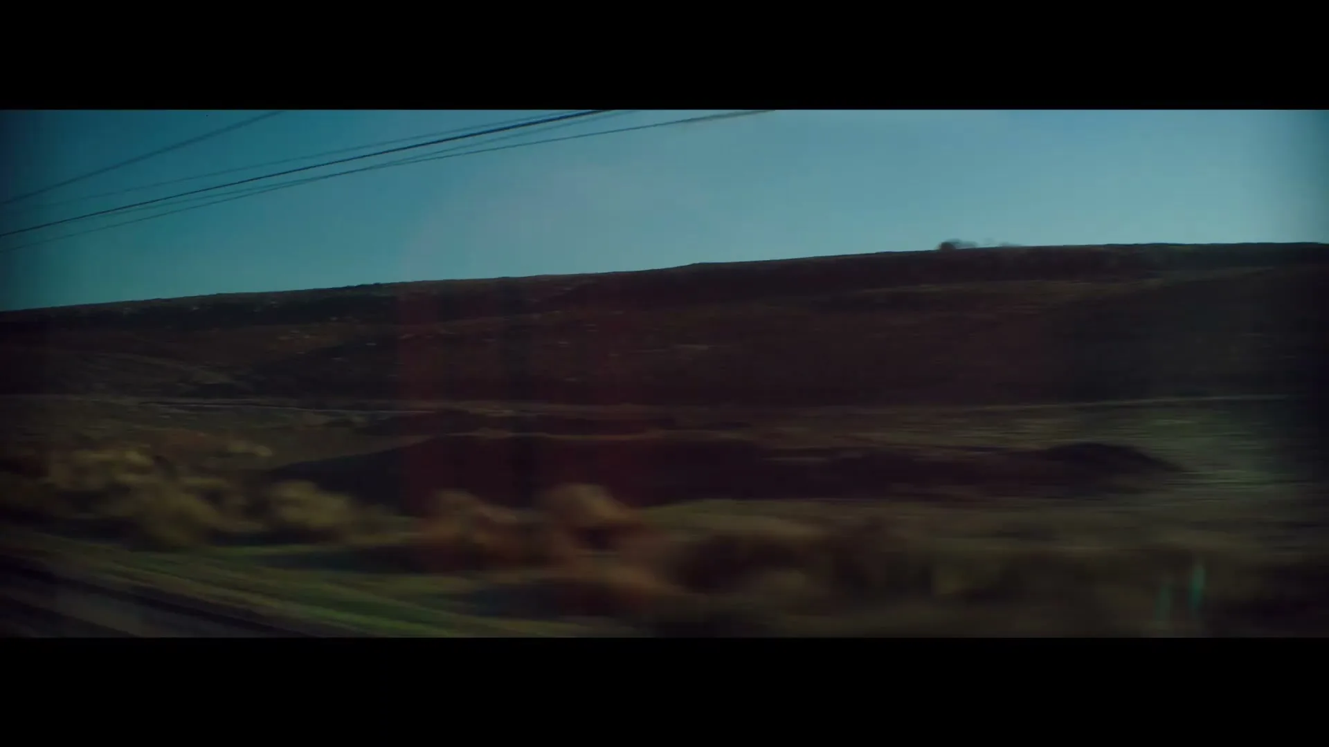 A blurry, wide shot from a moving train shows a vast, arid landscape with hills and a clear blue sky during the day.