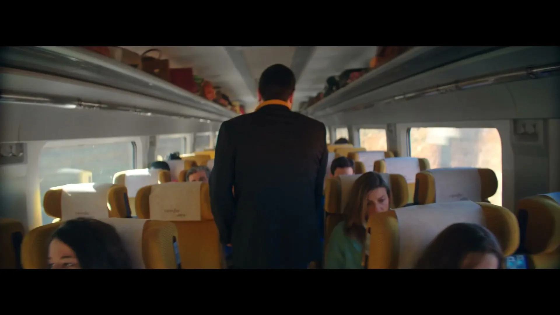 A man in a suit stands in the aisle of a moving train, looking forward, as passengers sit in their seats.