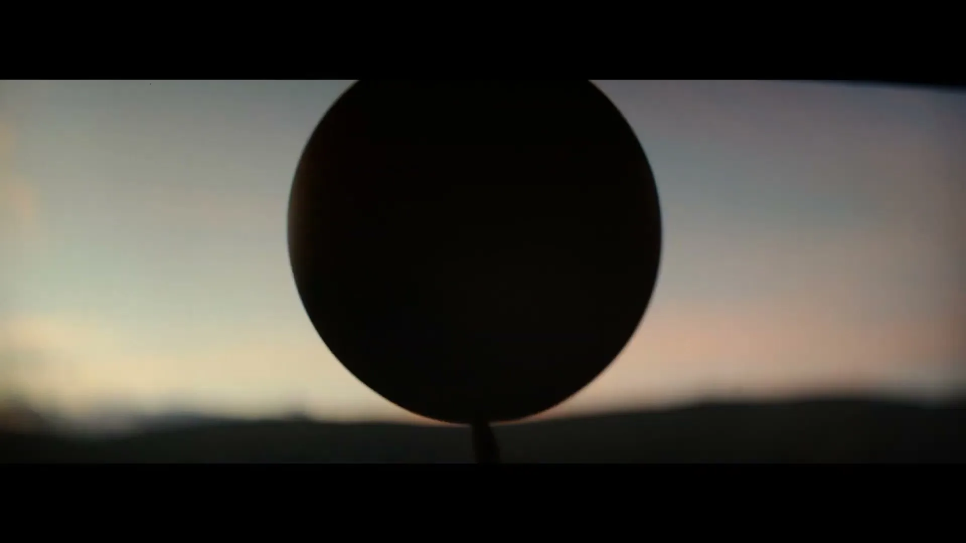 A dark, circular object, possibly a balloon, is silhouetted against a soft, colorful sky at dusk, with distant hills blurred in the background.