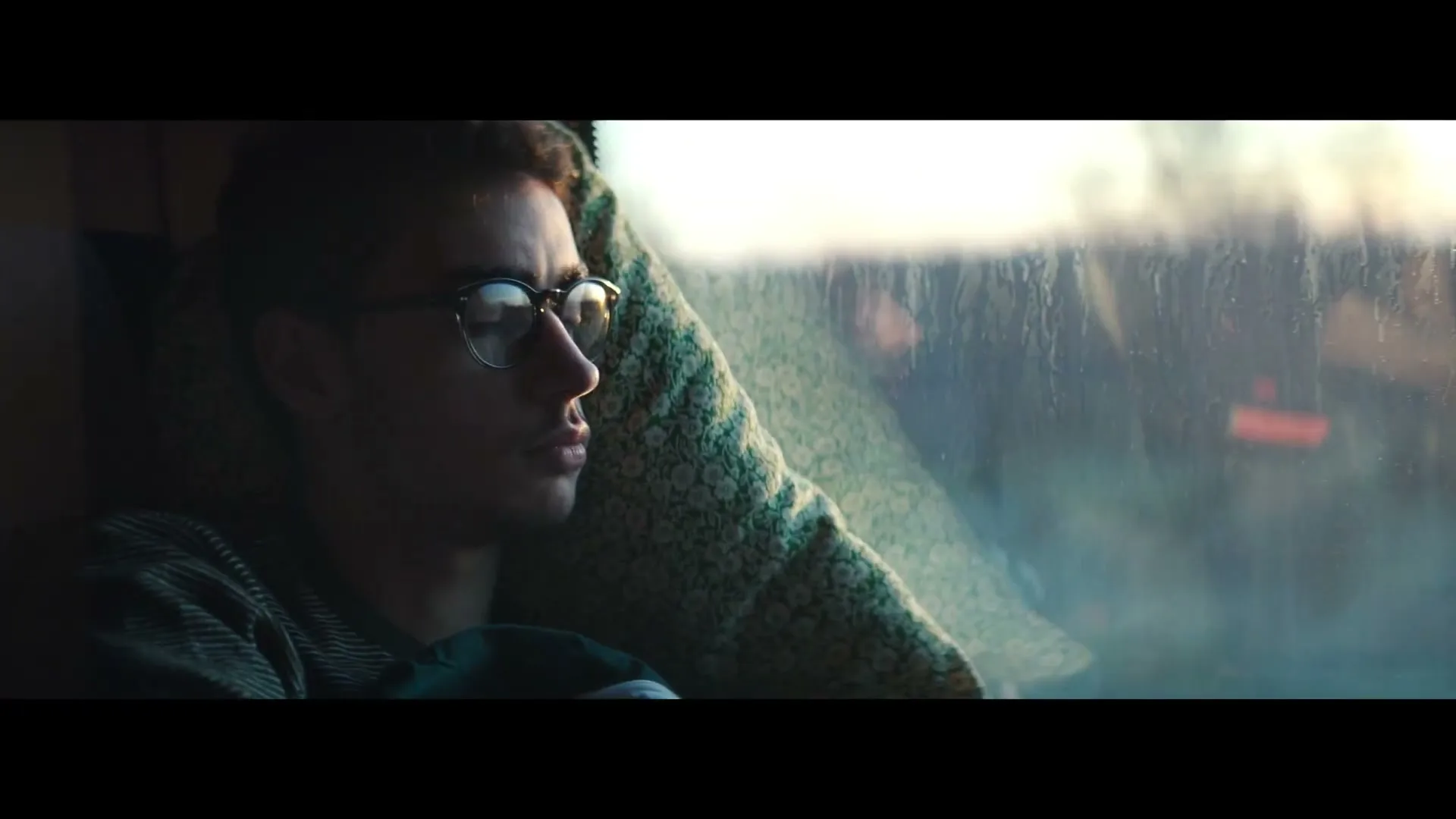 A young man with glasses rests his head on a pillow, looking out a train window as the landscape blurs past at dusk.