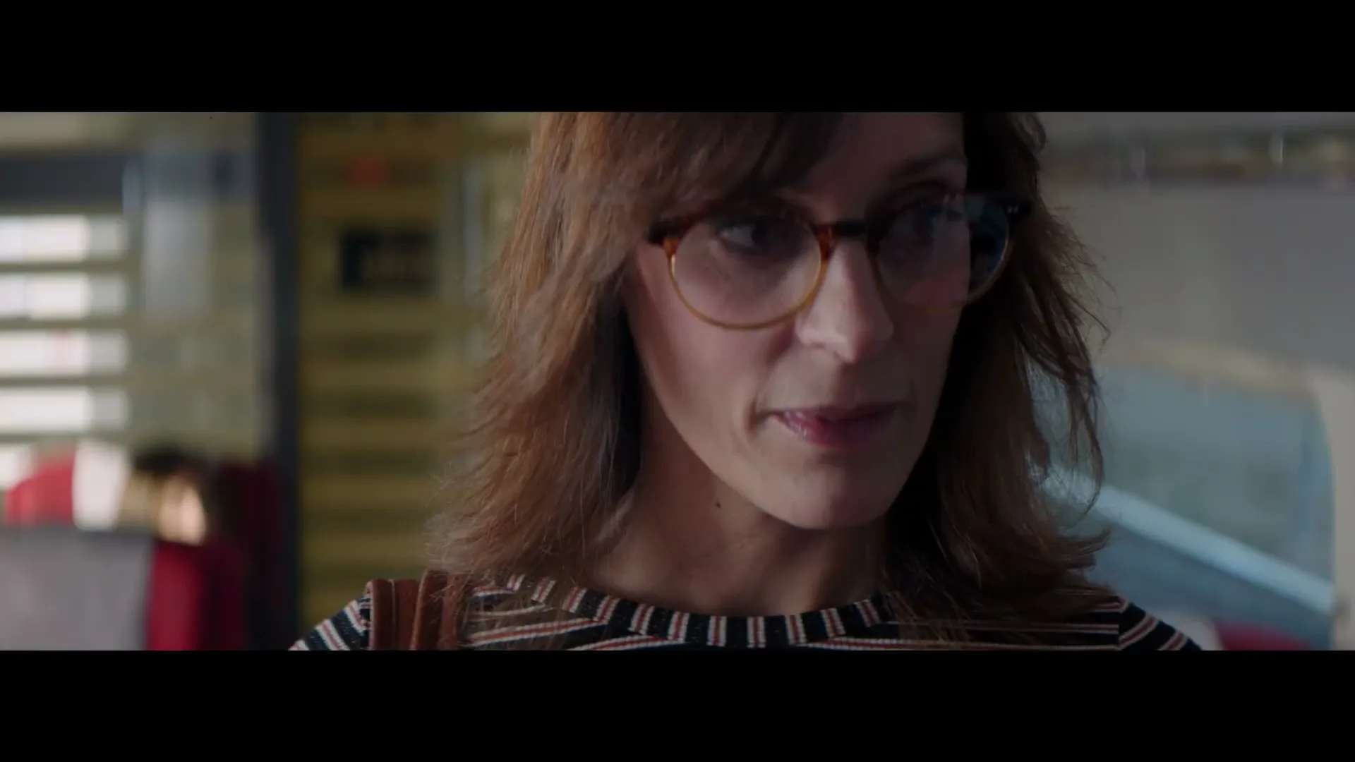 A woman with brown hair and glasses looks down, then up, with a slight smile in a brightly lit interior.