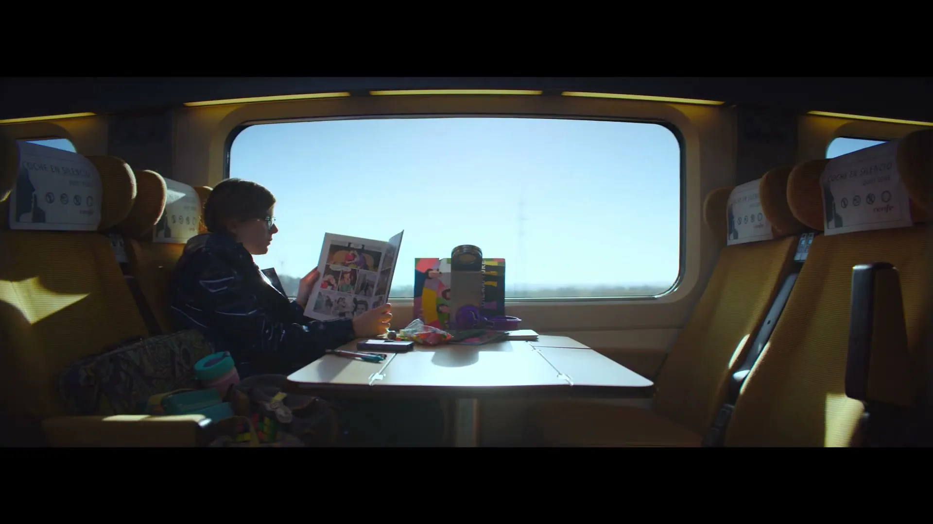 A young boy with short hair and glasses sits at a table on a train, reading a comic book, with sunlight streaming through the window.