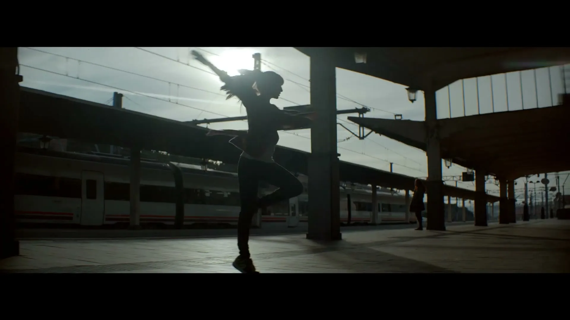 A silhouetted young woman with a ponytail dances gracefully on a train station platform, with a train visible in the background under a bright sky.