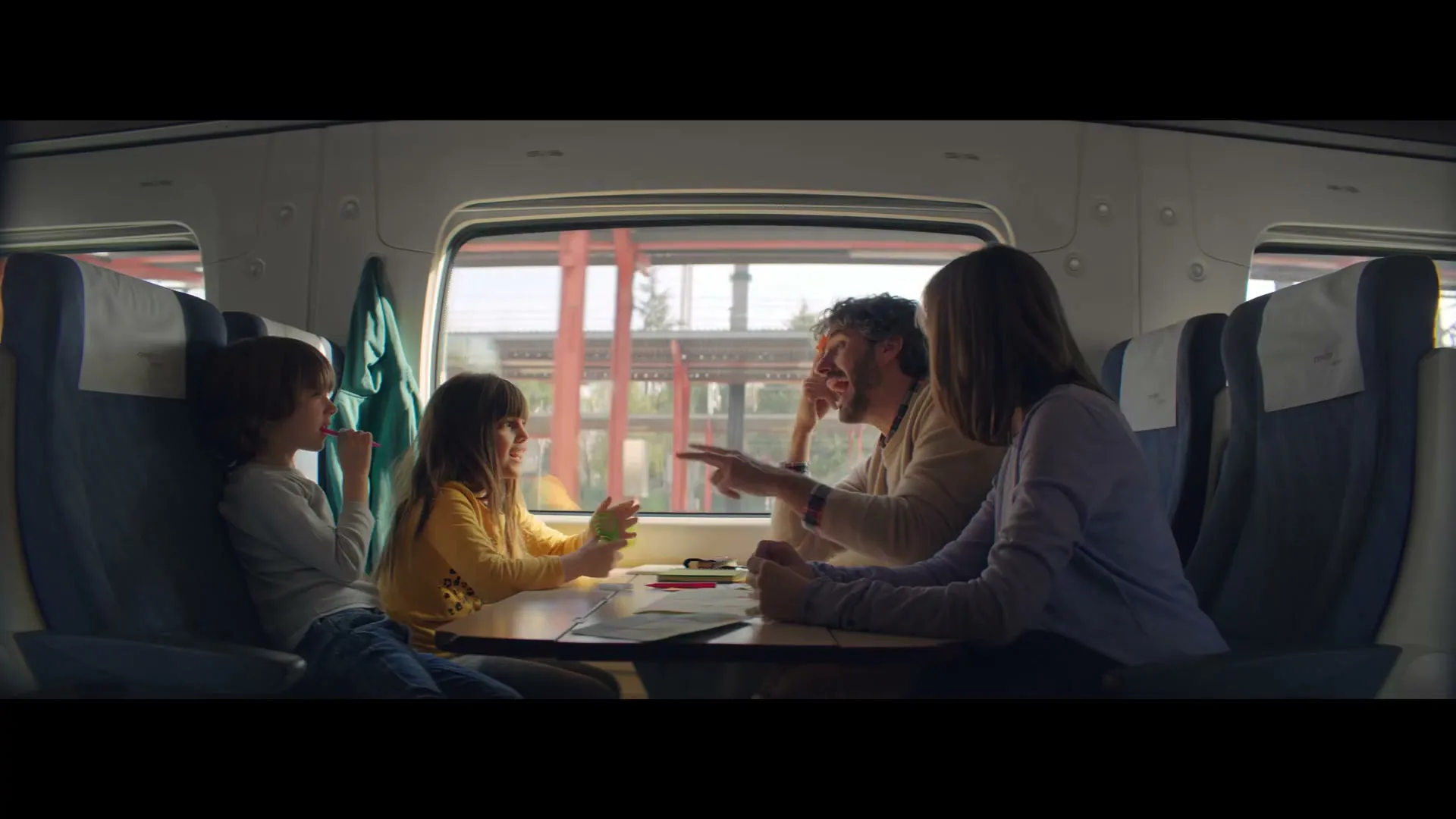 A family of four, two children and two adults, sit at a table inside a train, looking out the window and talking.