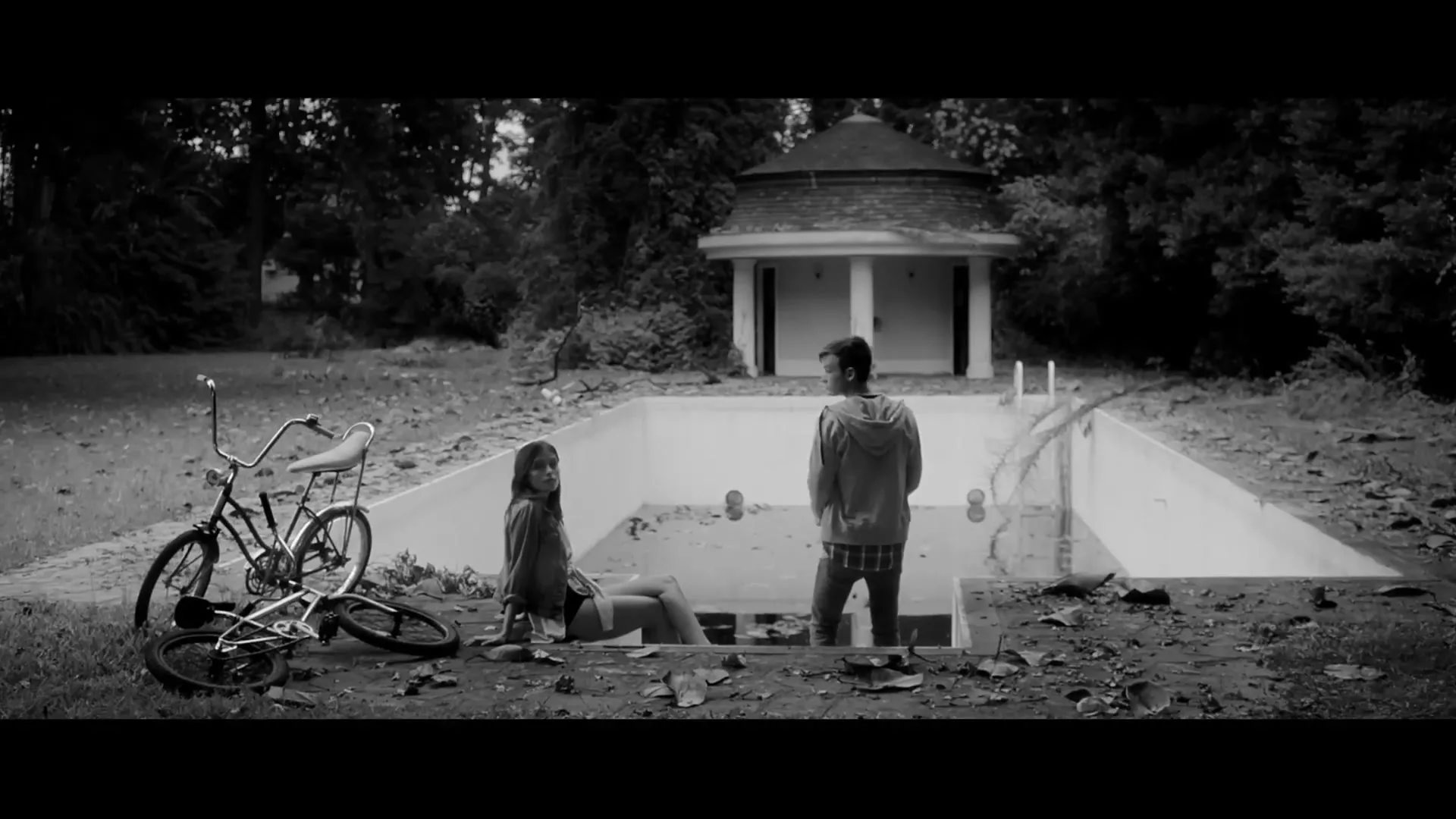 A young man and woman stand in an empty, dirty swimming pool in a dilapidated backyard with a small gazebo, in black and white.