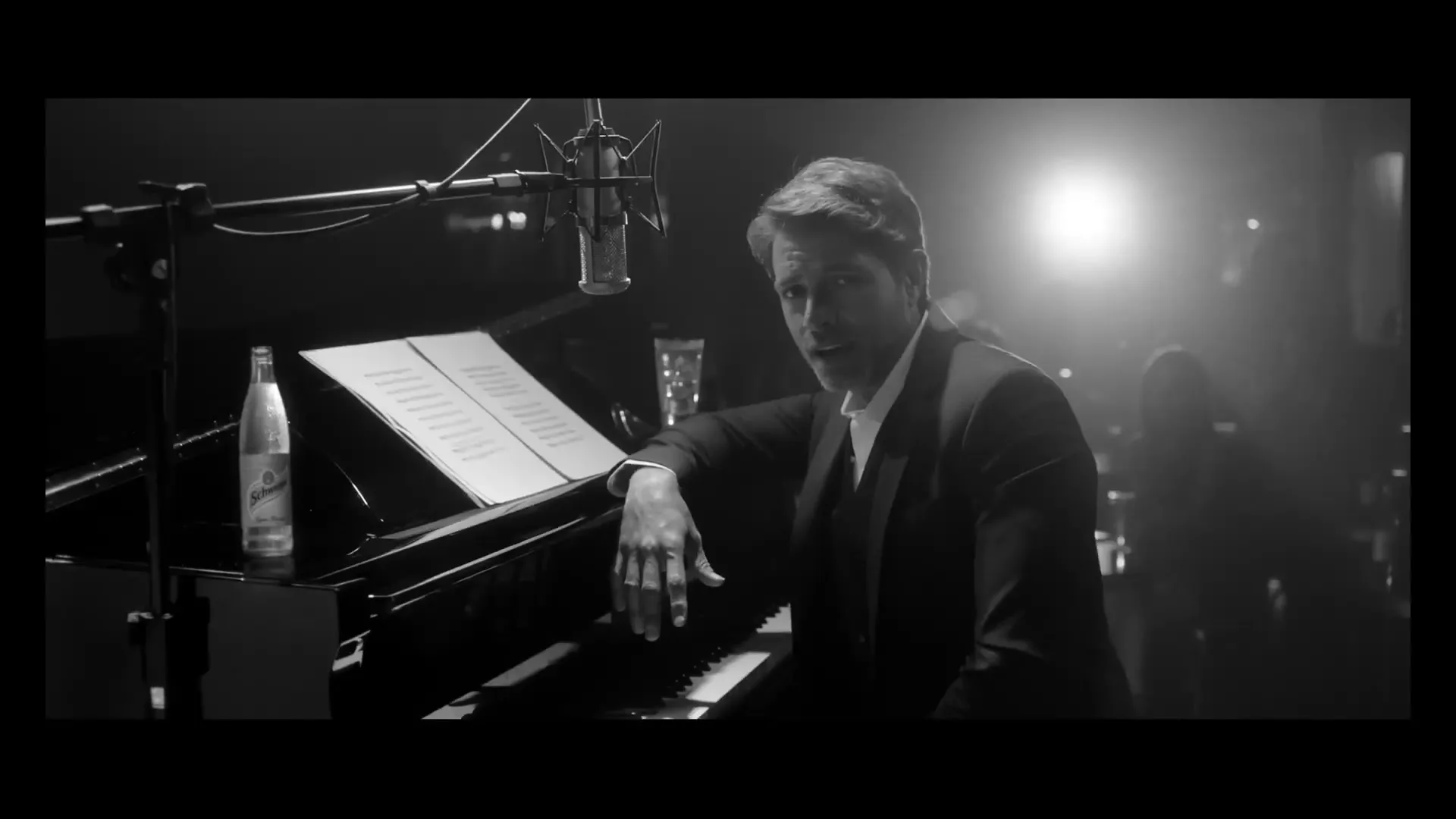 A man in a suit sits at a piano with sheet music and a microphone, looking directly at the camera in a dimly lit club.