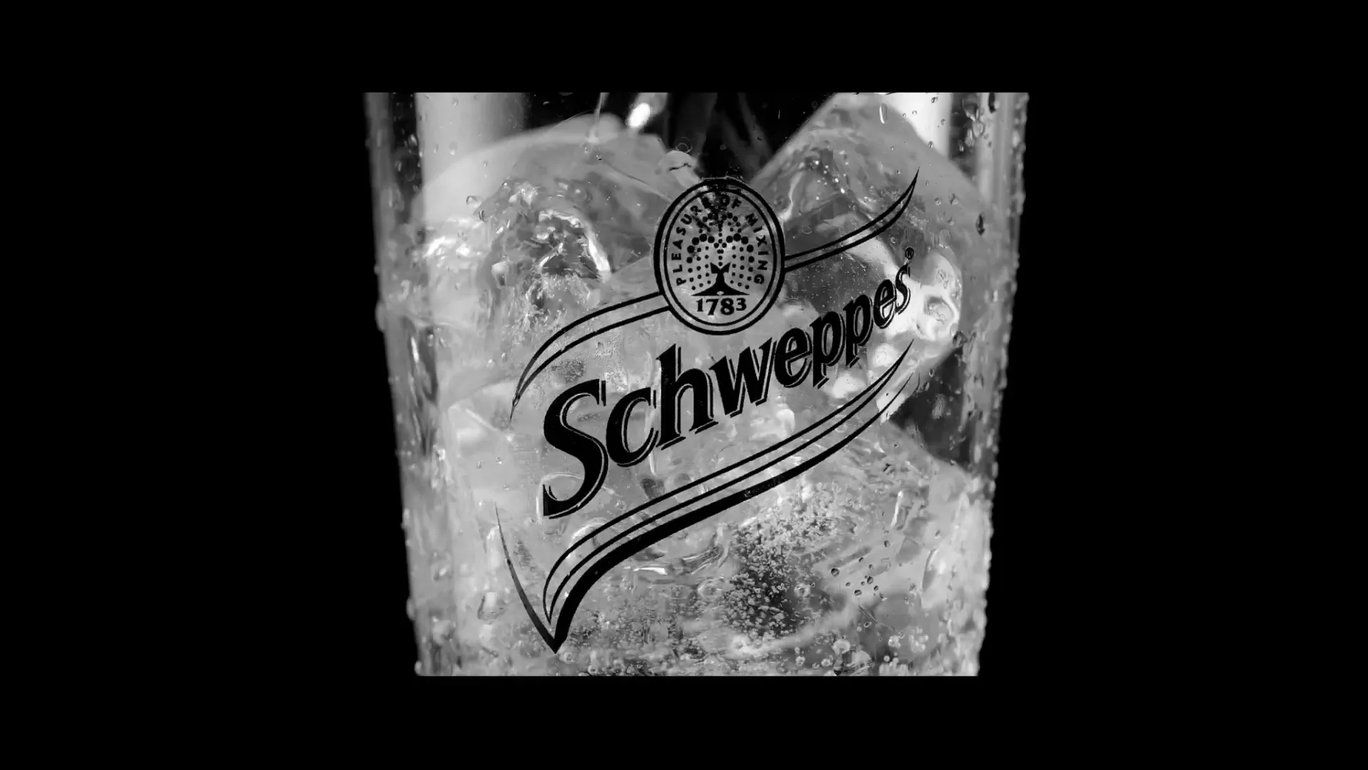 A close-up shot of a glass filled with ice and a Schweppes logo, with liquid being poured into it, against a black background.