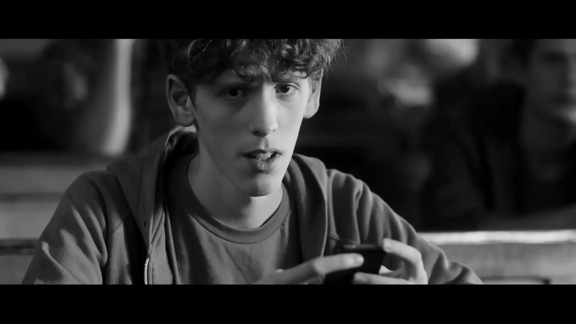 A young man with curly hair, wearing a hoodie, looks up from his phone with a surprised expression in a dimly lit interior.