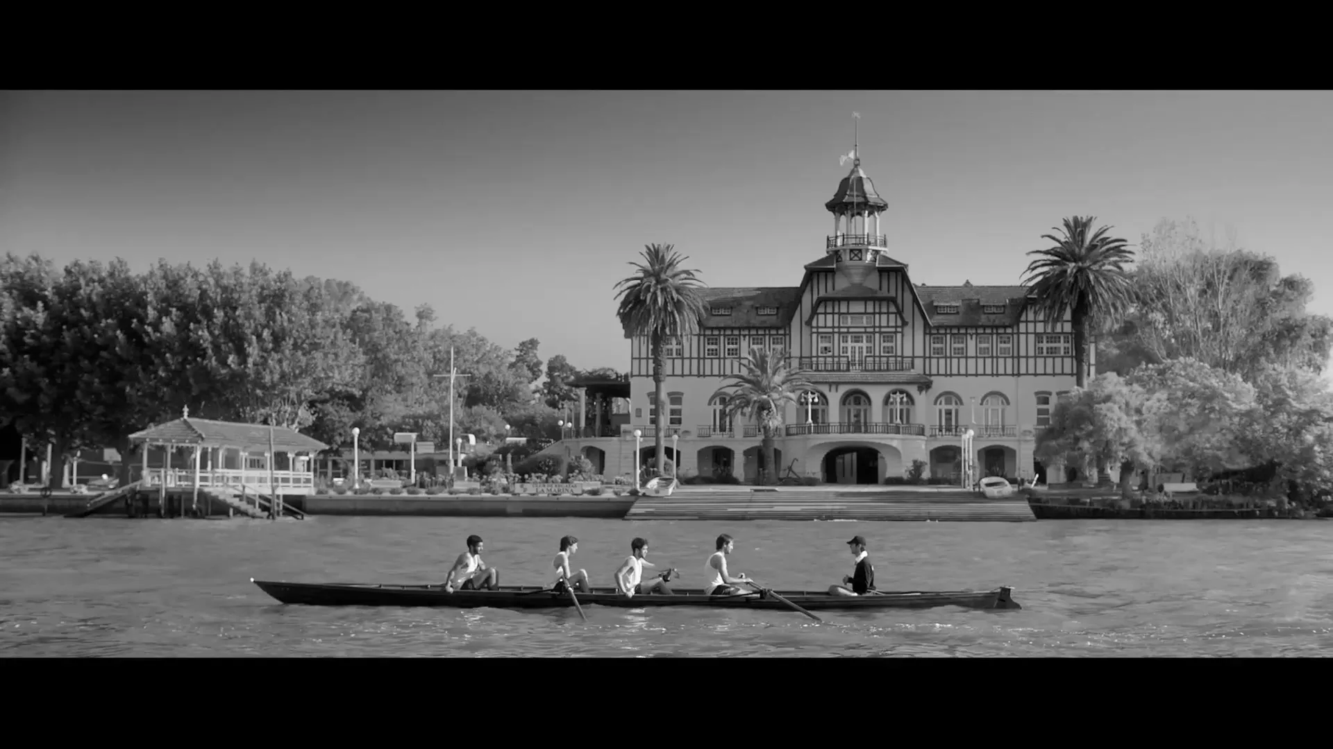 Five men in a rowing boat on a river, with a large, ornate building and trees on the bank in the background, in black and white.
