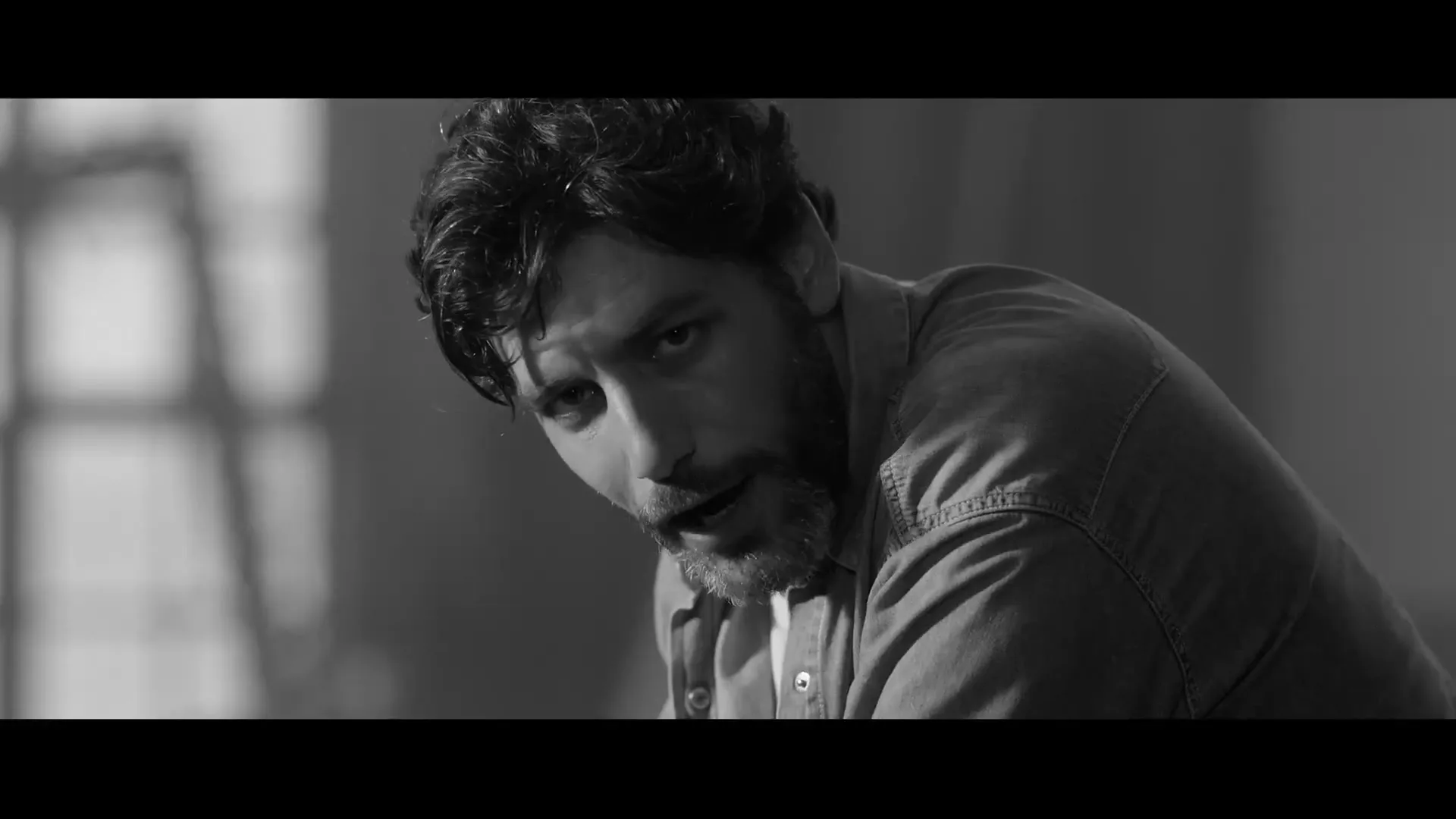 A man with dark hair and a beard, wearing a denim shirt, looks intently at the camera in a dimly lit interior.