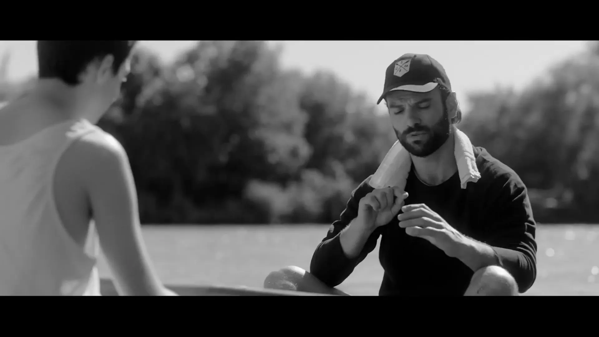 A man in a cap and towel around his neck sits in a boat, talking to another person, on a sunny day.
